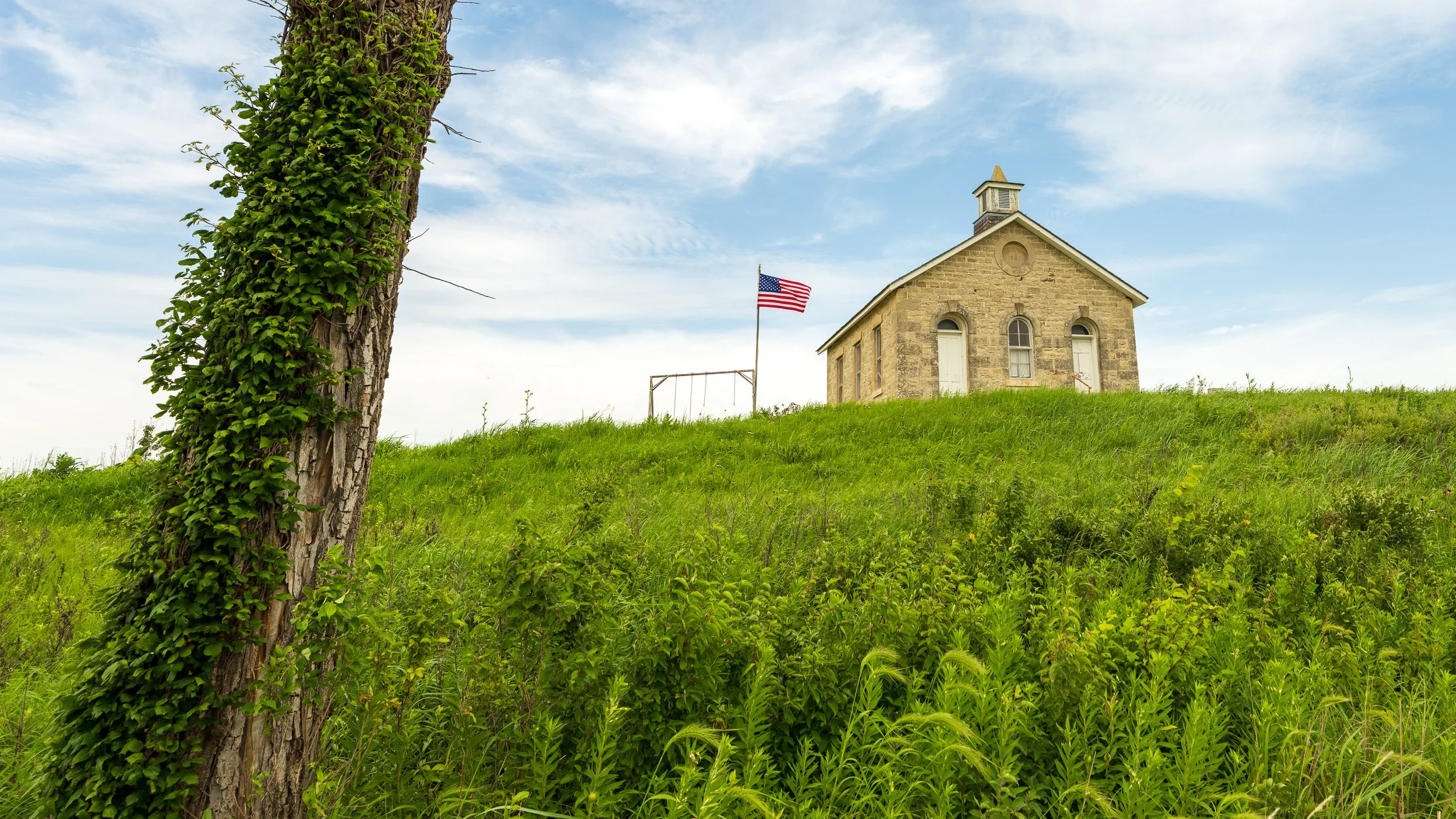 A small stone church with a bell tower, surrounded by green grass and trees, under a partly cloudy sky, with an American flag flying nearby.