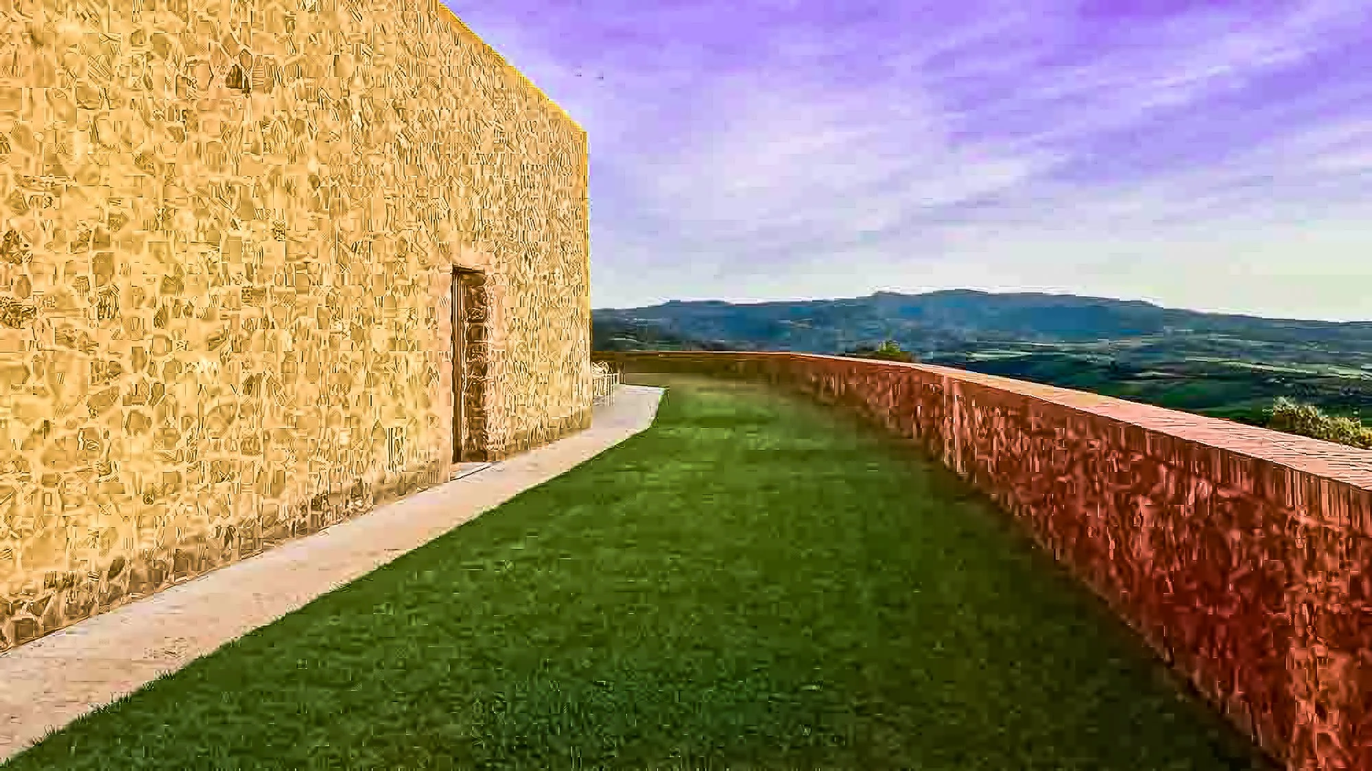 Photo of a stone building with a door, a green grass lawn, a brick wall, and a distant mountainscape under a purple-tinged sky.