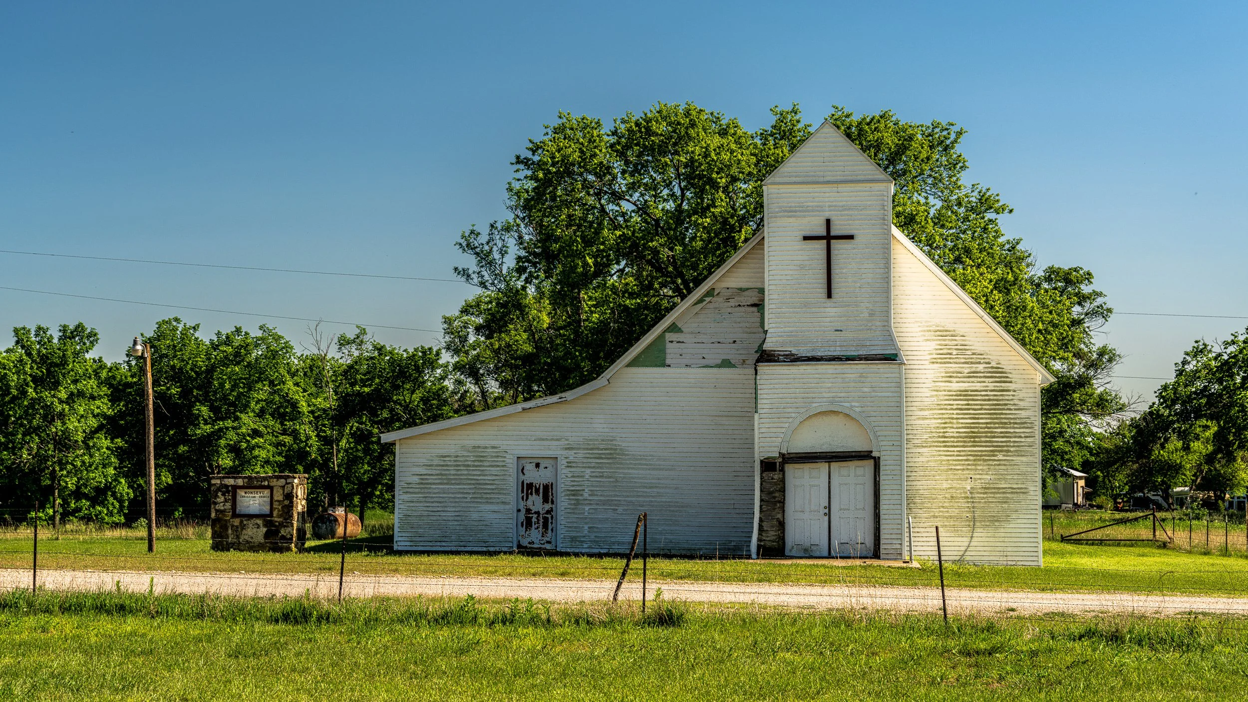 A white wooden church with a cross on its front, surrounded by green trees and grass, under a clear blue sky.