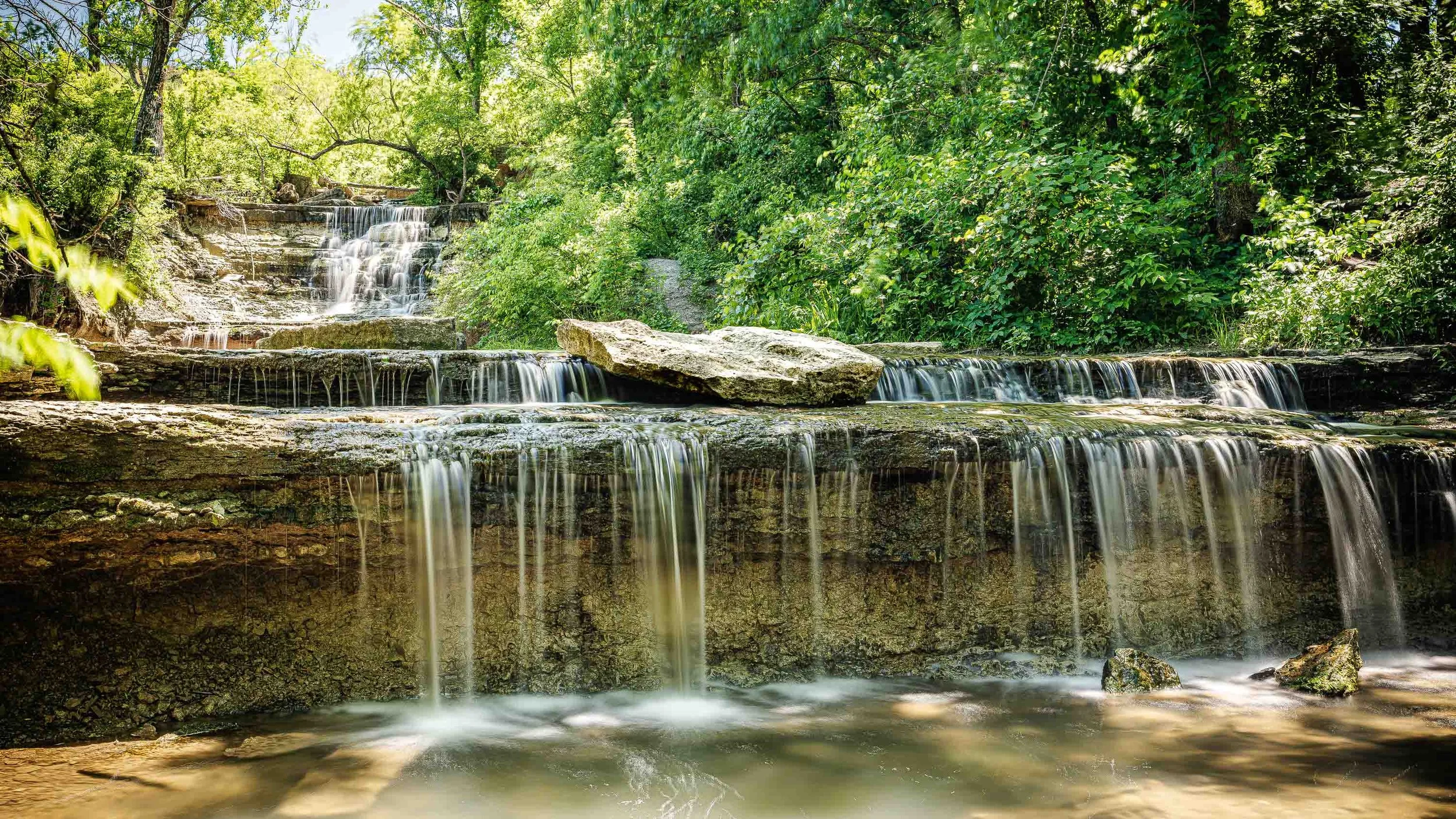 A multi-tiered waterfall flowing over rocks surrounded by lush green trees and foliage in a forest setting.