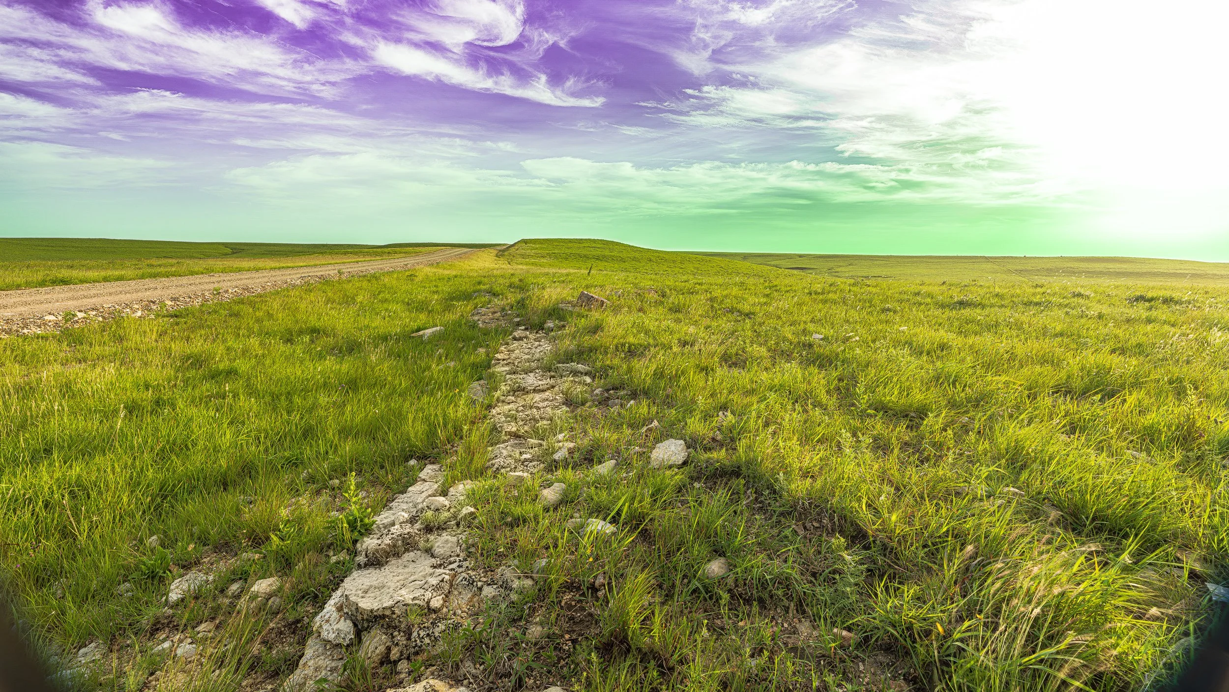 Open grassy field with a dirt and stone path leading into the distance, green hills, and a sky with scattered clouds and a bright sun.