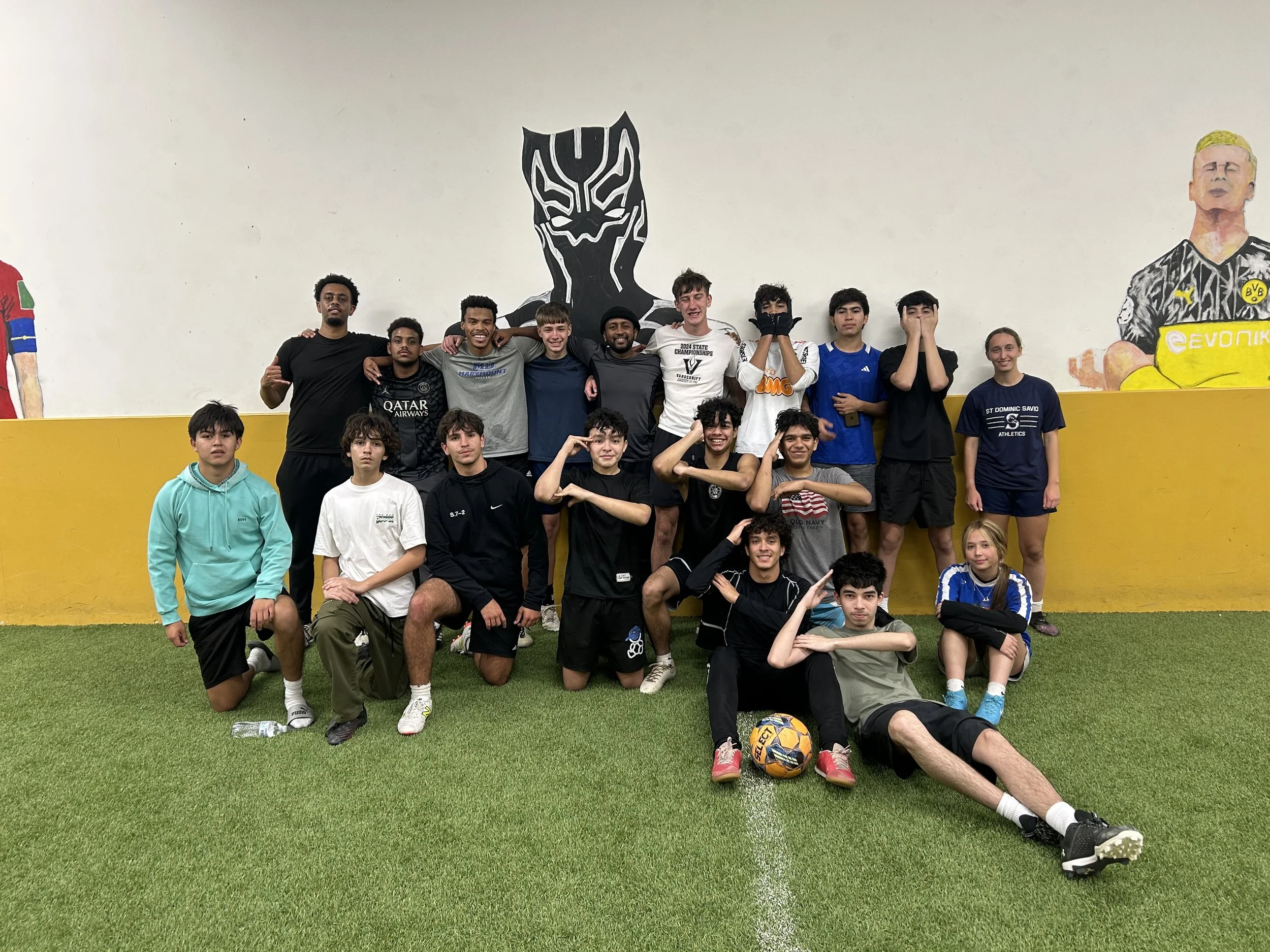 A group of young boys and teenagers posing for a photo inside a sports facility with a large mural of a soccer player and a person with a painted mask on the wall behind them. Some players are kneeling, some standing, and they are all smiling or making gestures. A soccer ball and a water bottle are on the ground in front of them.
