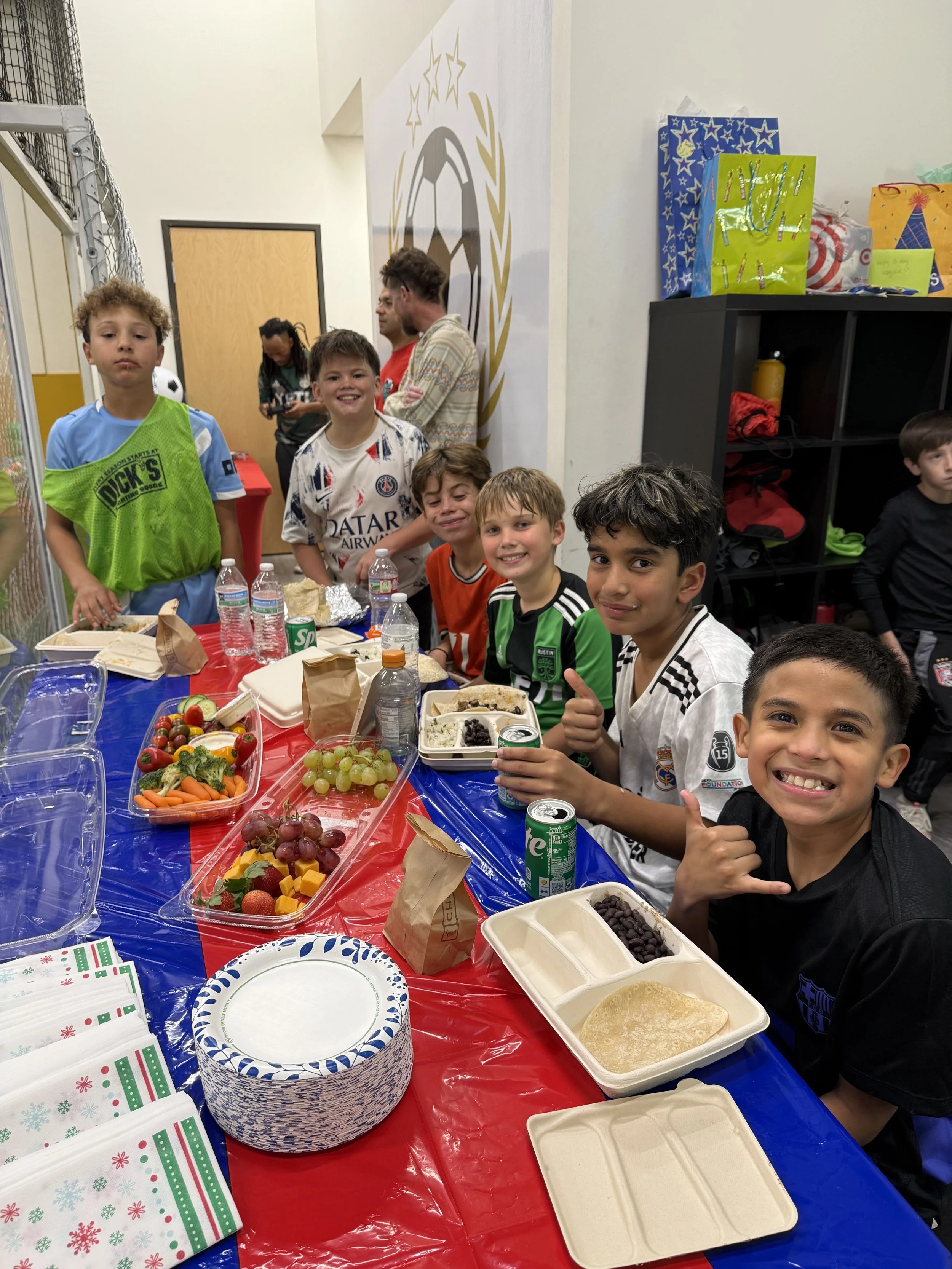 A group of young kids standing on a football field under a partly cloudy sky, some wearing sports uniforms and holding a soccer ball.