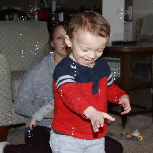 Child smiling and popping bubbles during a speech therapy activity with his mother.