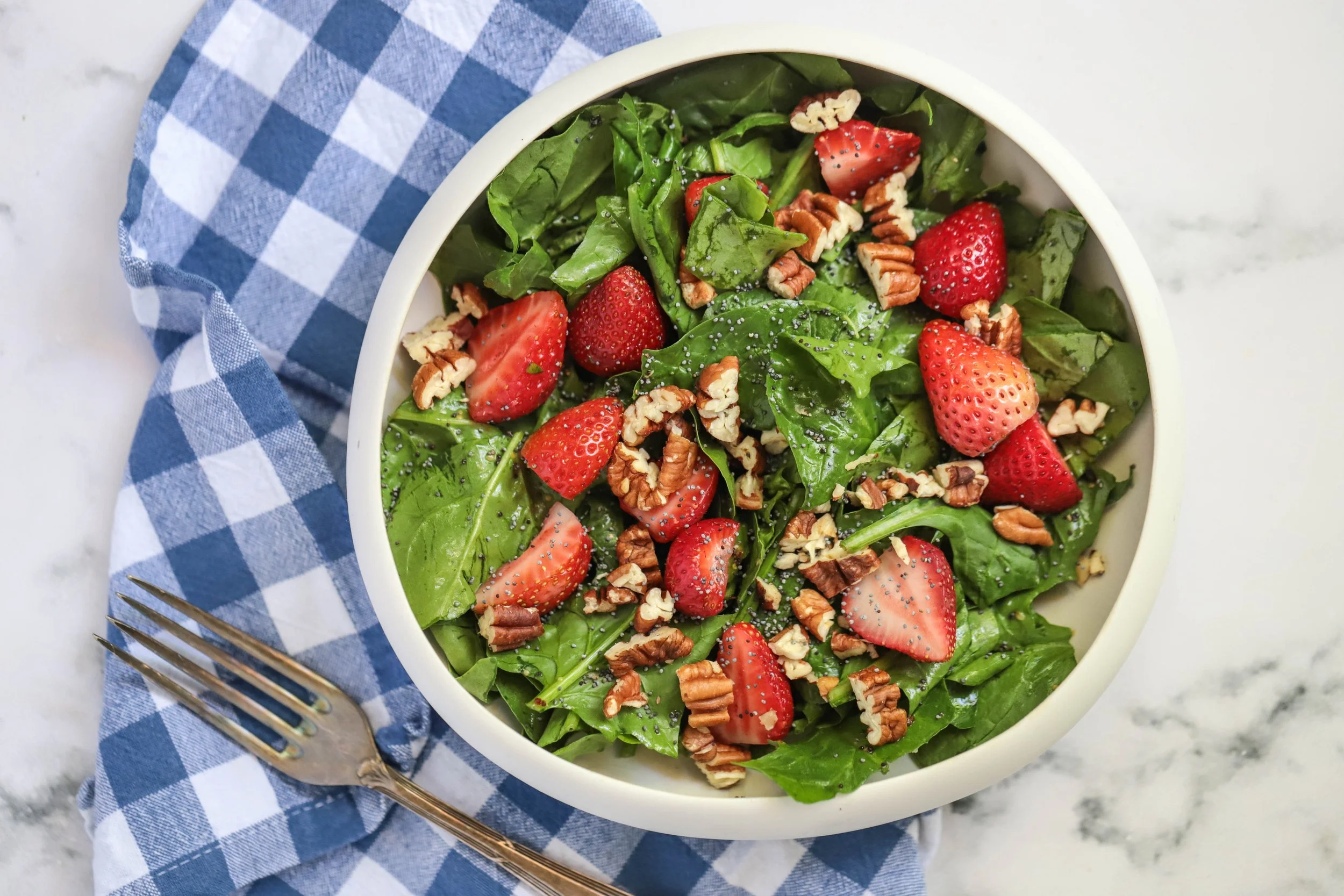 Photograph of strawberry spinach pecan salad in a white bowl next to a black and white table cloth with a silver fork on a marble countertop shot from above