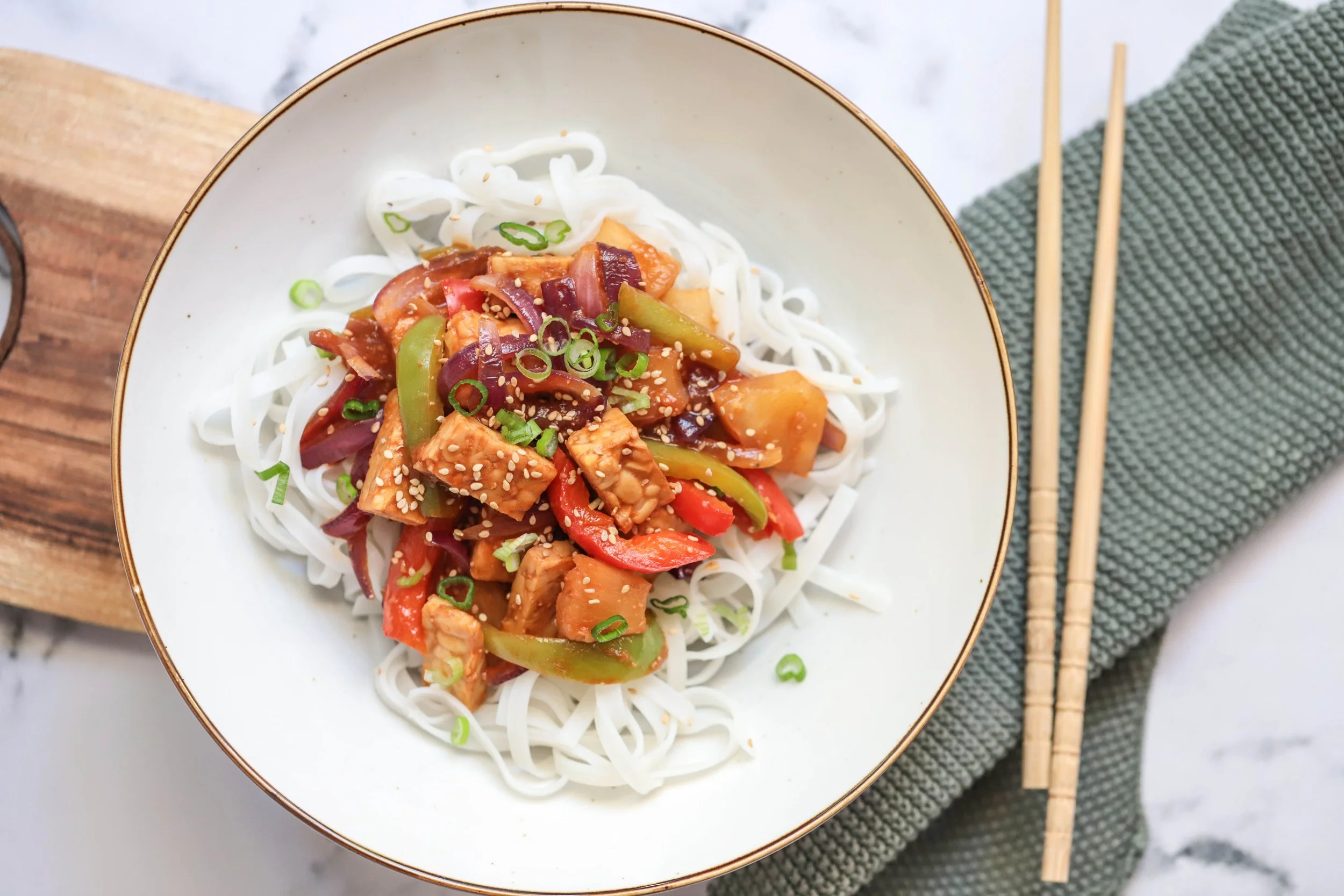 Sweet and sour tempeh on rice noodles in a white bowl with gold rim sitting on a wooden cutting board with a tea towel next to it and chop sticks on a marble countertop