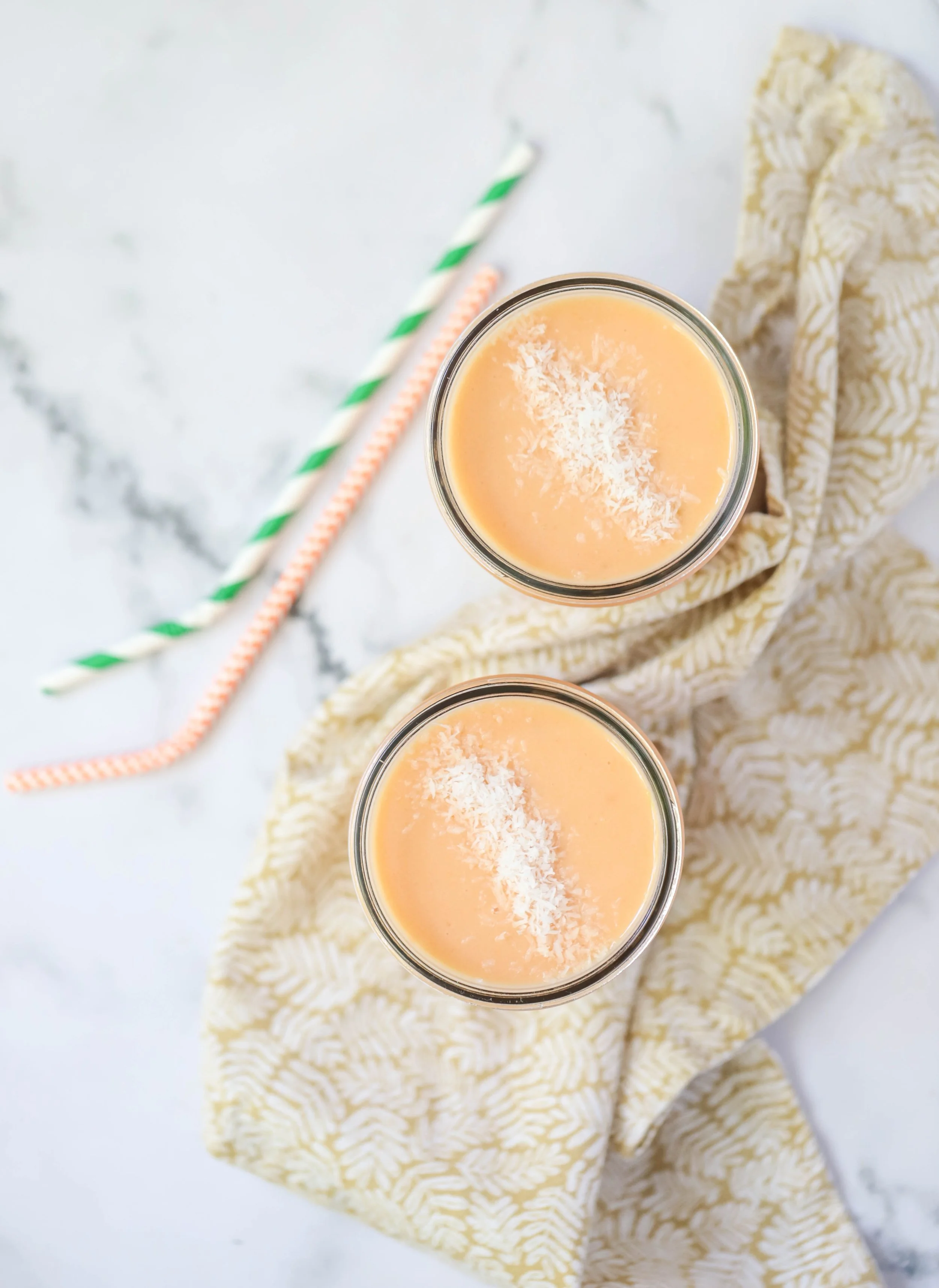 Two papaya mango smoothies in glass jars with paper straws on a gold and beige napkin on a marble countertop
