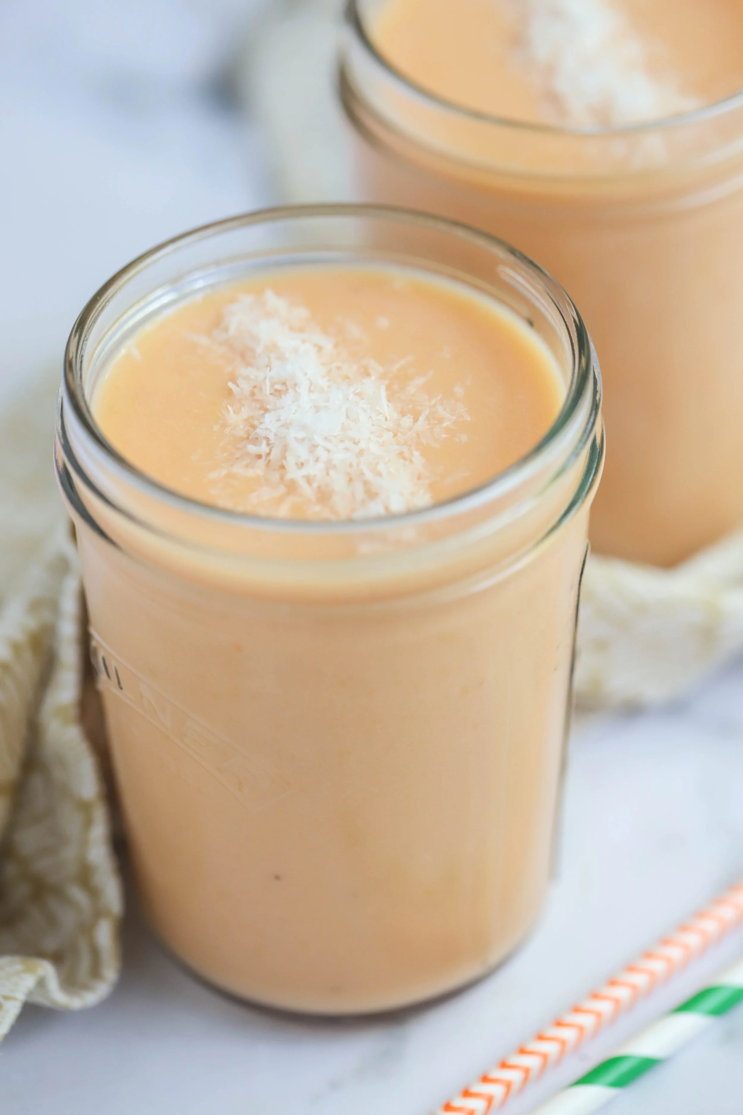 Side angle of Two papaya mango smoothies in glass jars with paper straws on a gold and beige napkin on a marble countertop