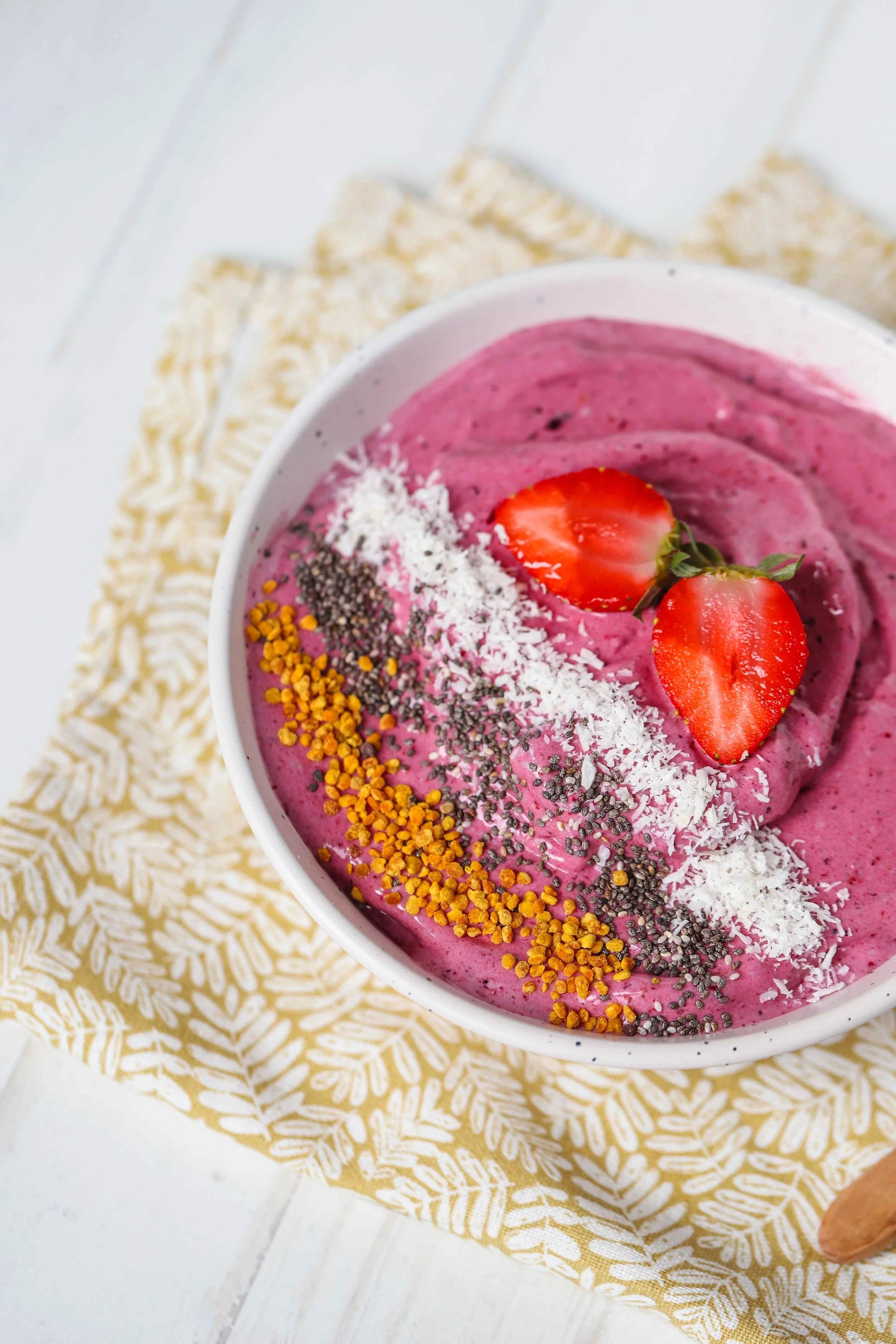 Close up of a red berry protein smoothie bowl with wooden spoon in it photographed from above on a leaf patterned napkin on a white countertop