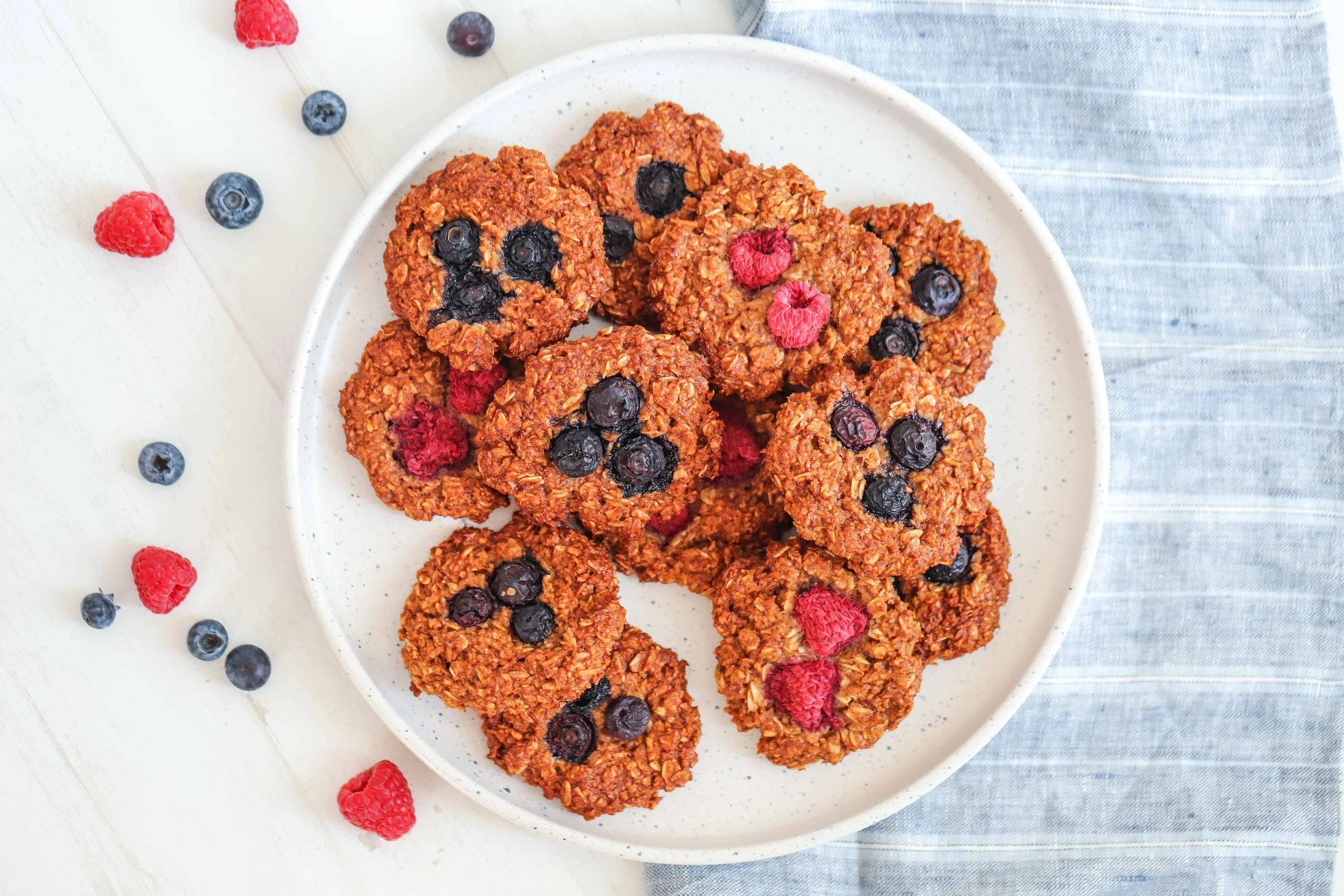 9 Oat Almond Meal Breakfast Cookies with Blueberries and Raspberries as garnish on a white plate next to a blue linen tablecloth on a white countertop