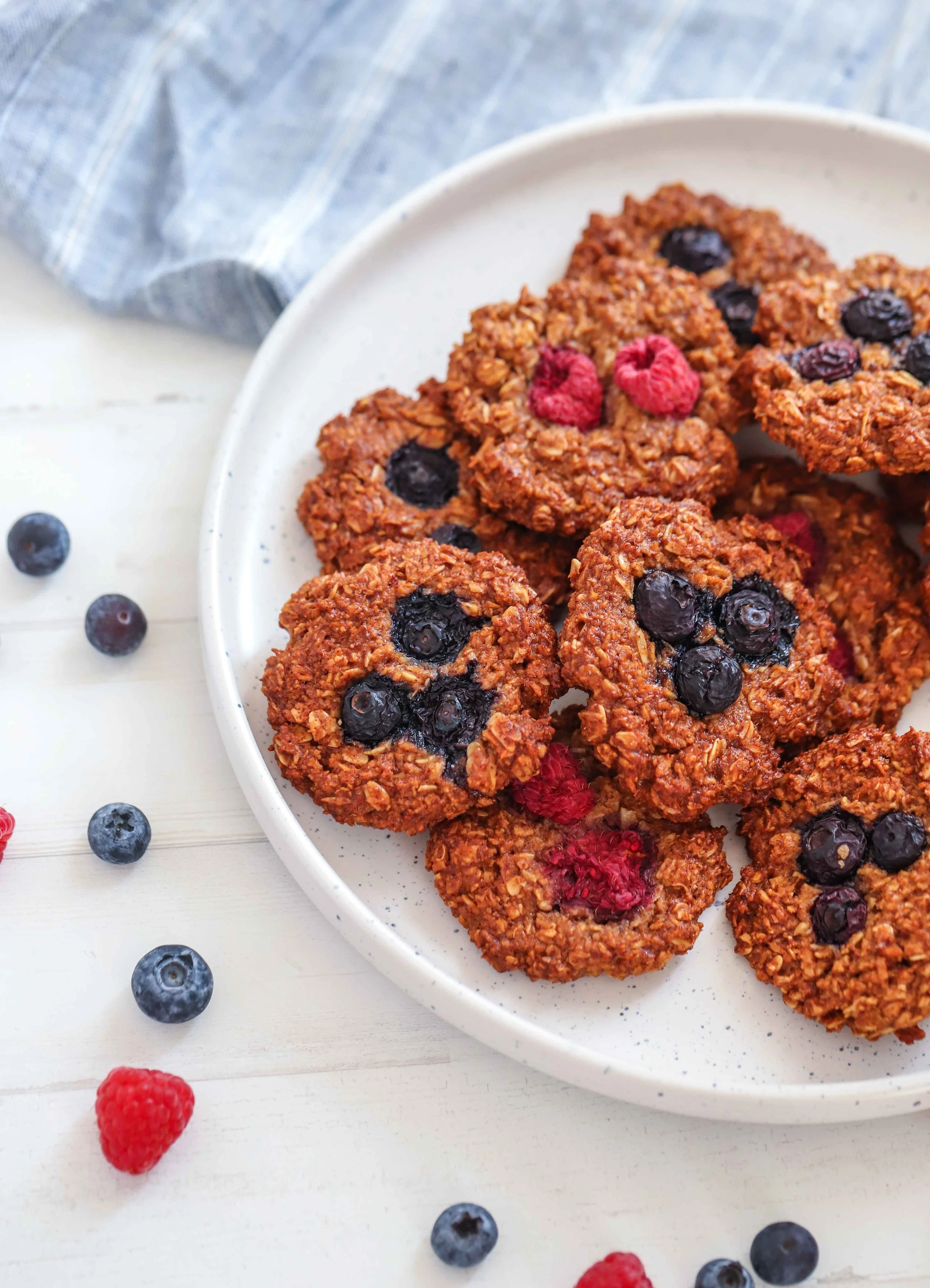 Close up of Oat Almond Meal Breakfast Cookies with Blueberries and Raspberries as garnish on a white plate next to a blue linen tablecloth on a white countertop