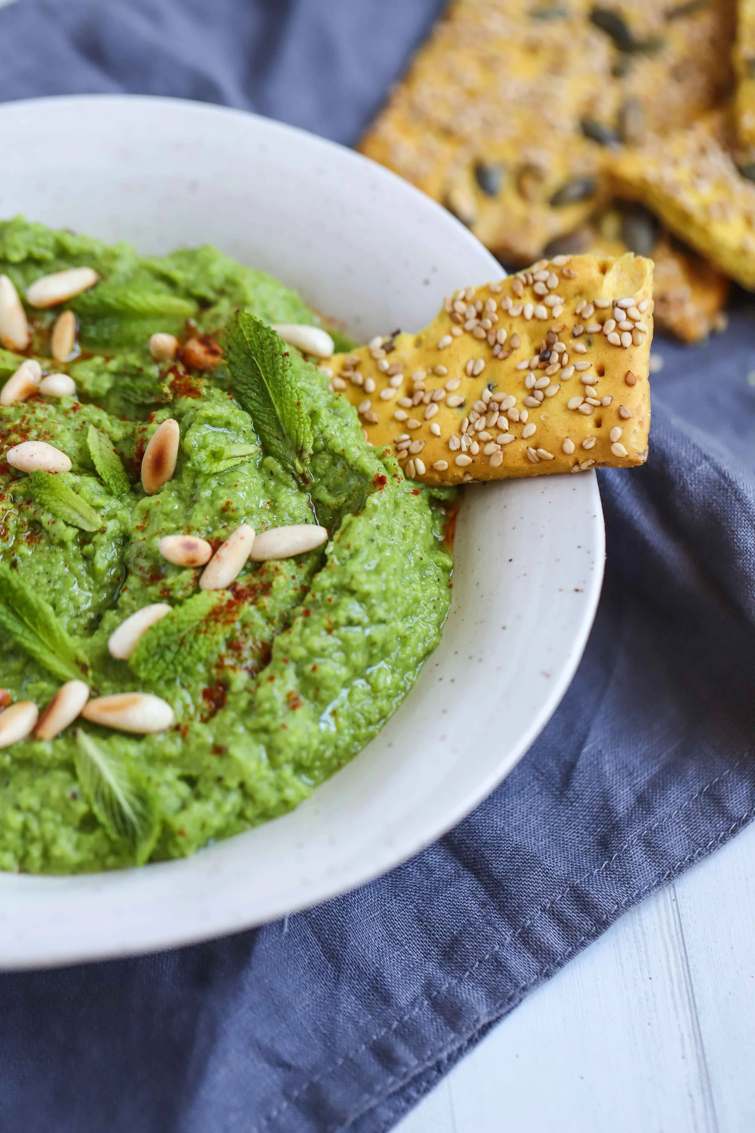 Mint avovado green pea dip topped with roasted pine nuts in a white bowl on a blue cloth next to seed crackers