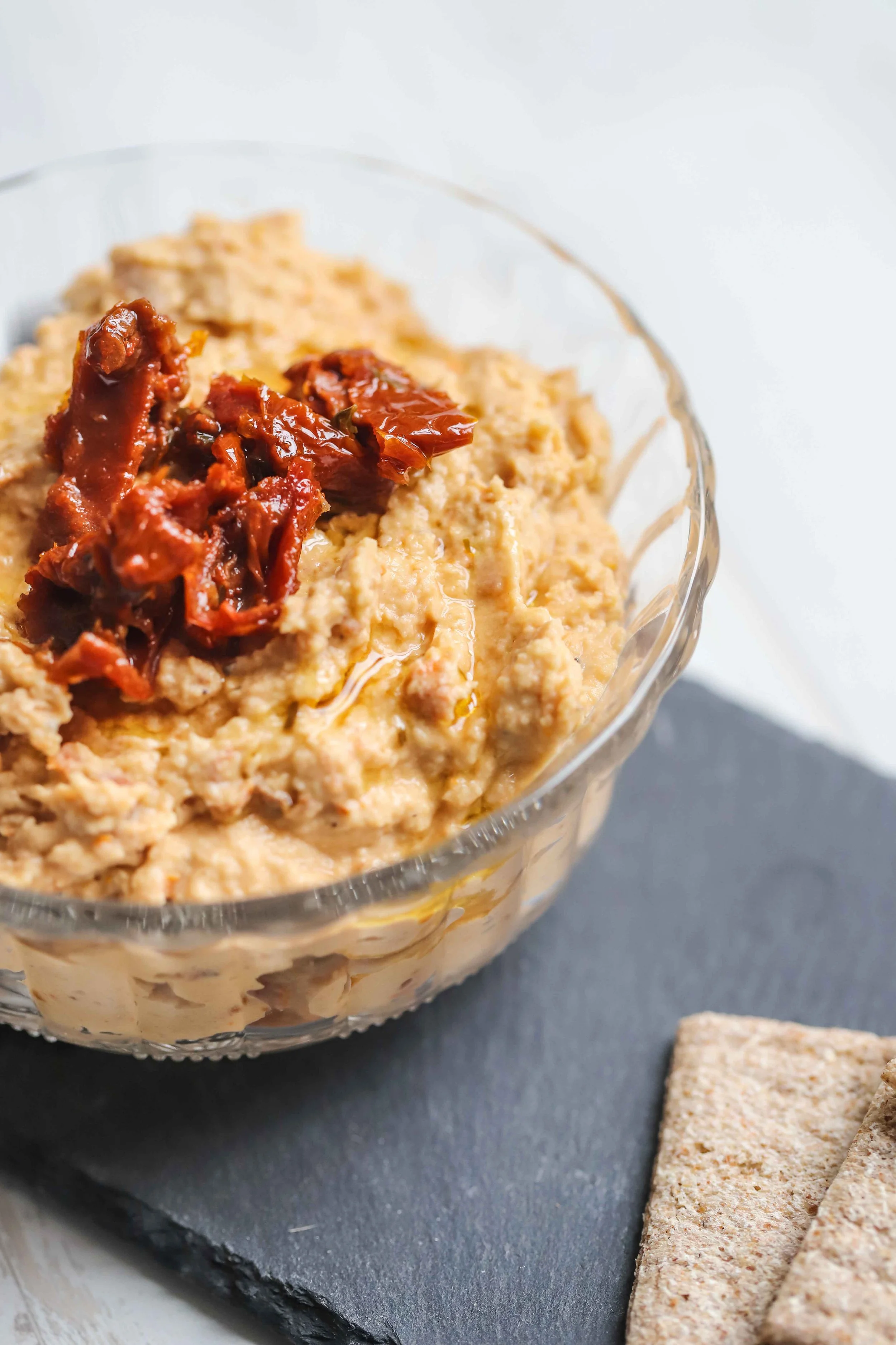Sundried tomato hummus in a glass bowl with crackers on a grey platter