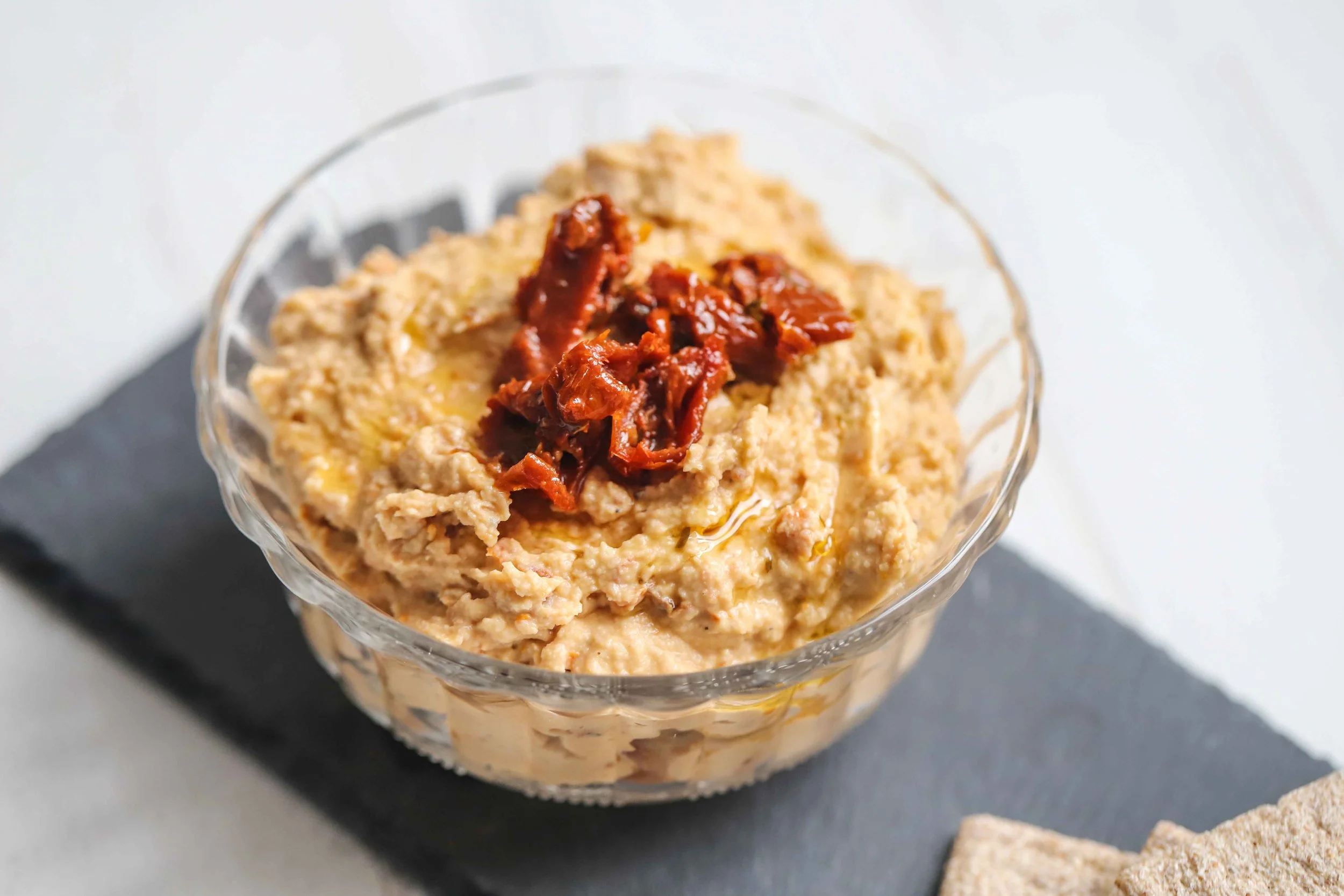Sundried tomato hummus in a glass bowl with crackers on a grey platter