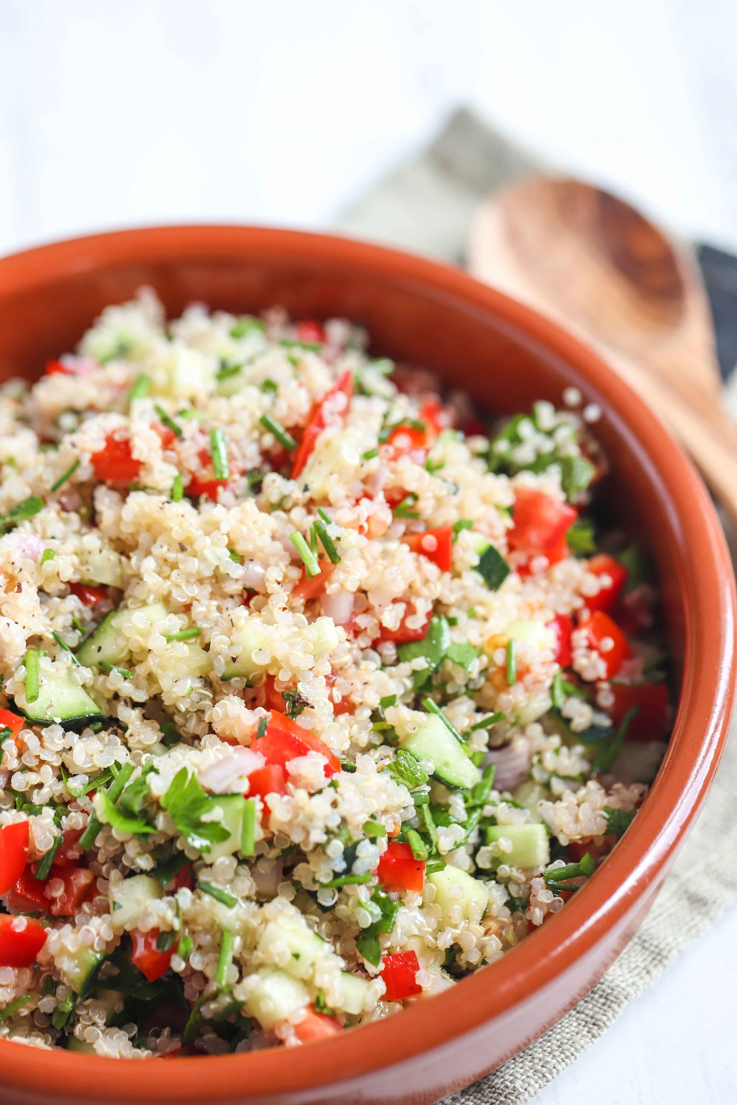 Quinoa tabbouleh in a red bowl on a linen napkin with a black stripe next to a wooden spoon