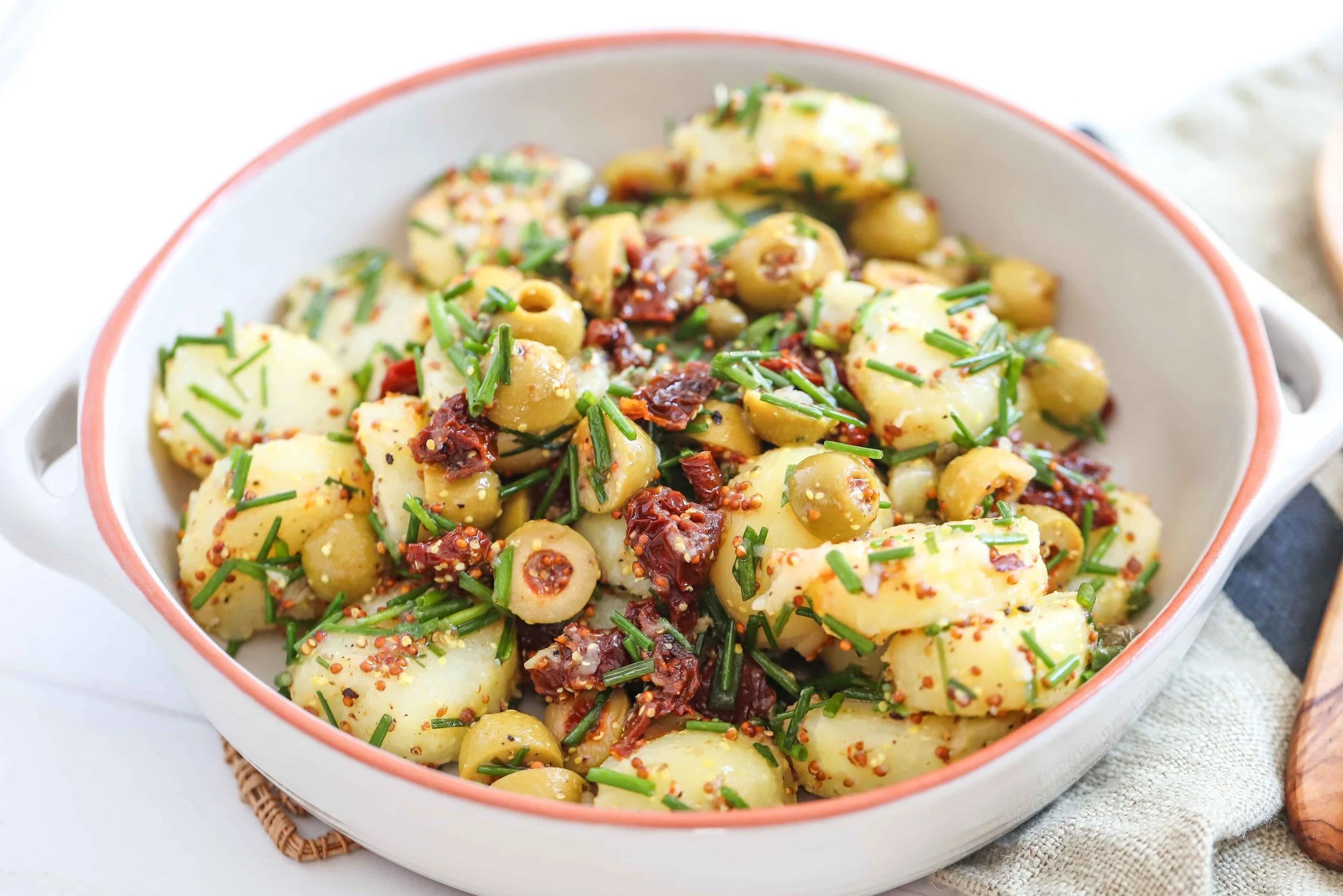 Sundried tomato potato salad in a white casserole dish with red rim next to a linen napkin with wooden serving utensils next to it