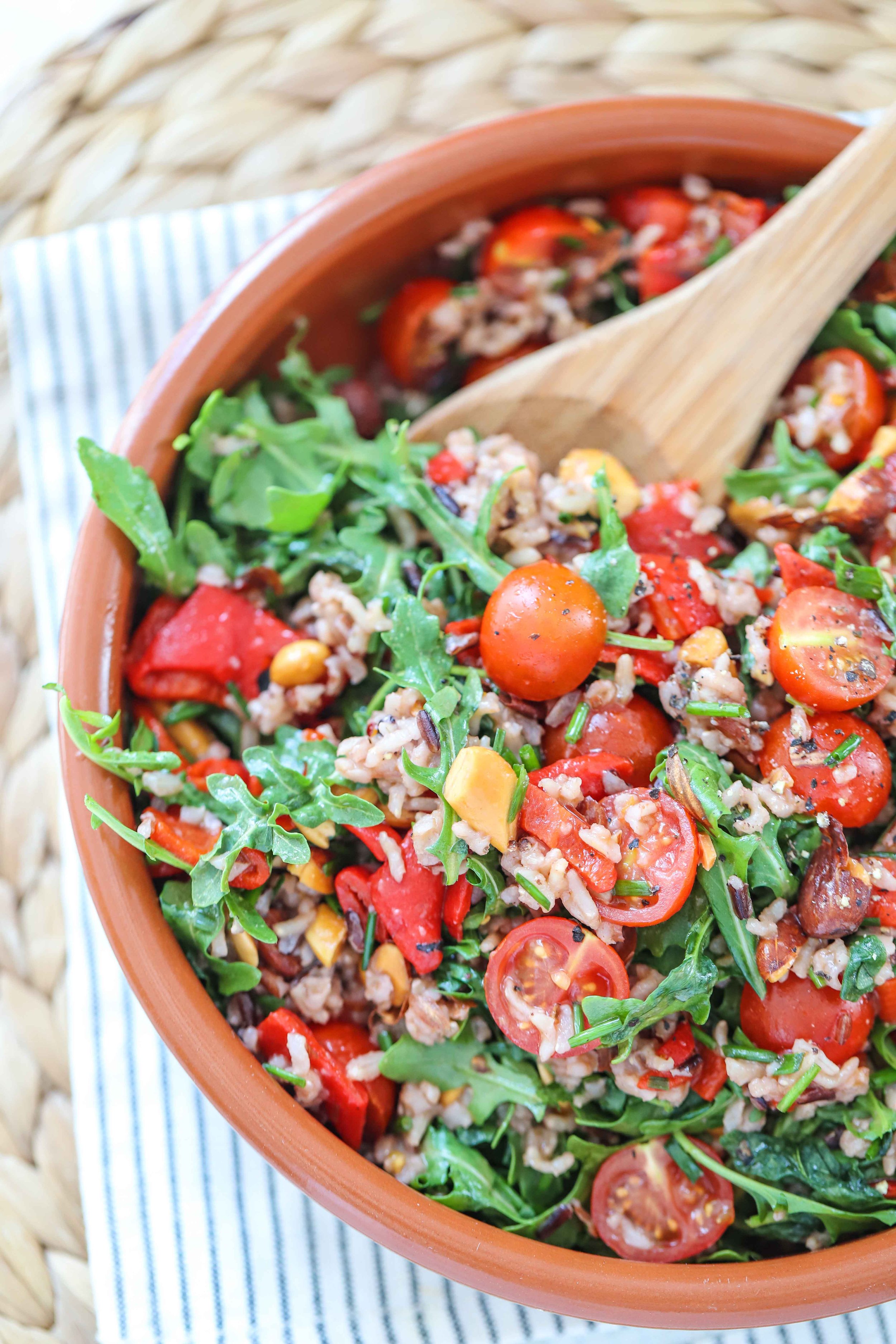 Tomato rocket wild rice salad with balsamic vinaigrette in an orange bowl on a blue and white napkin next to a wooden spoon with blue handle on a white countertop