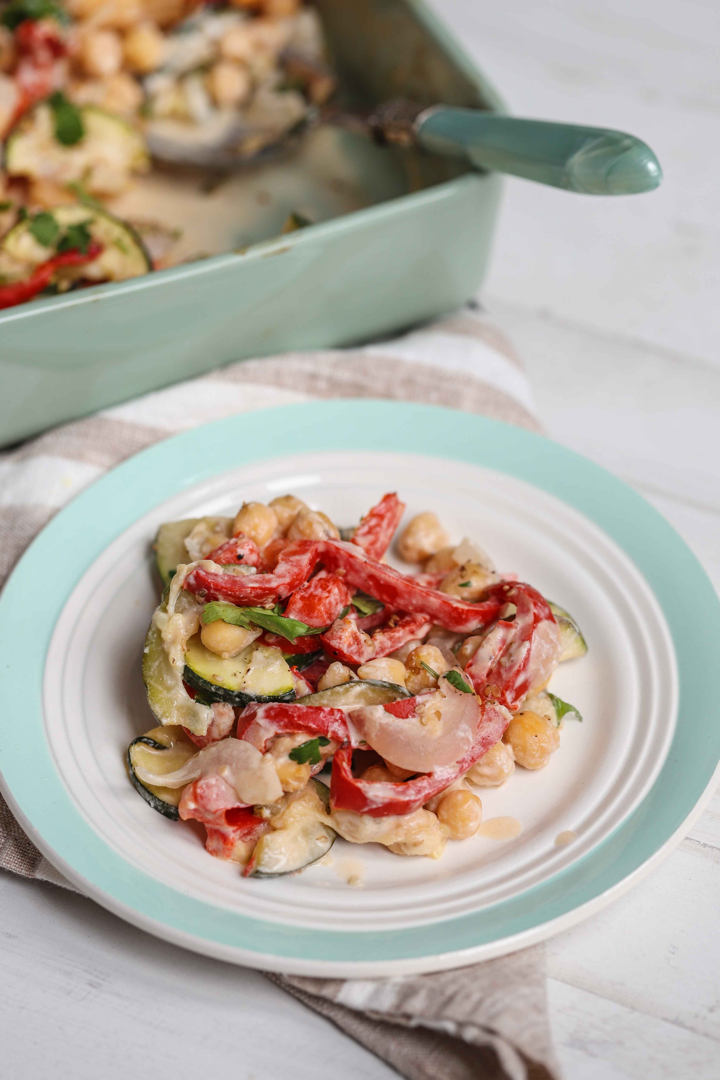 Baked vegetables with tahini on a plate next to the rest of it in a seafoam green pan with a seafoam green serving spoon in it on a linen napkin with white stripes on a white countertop