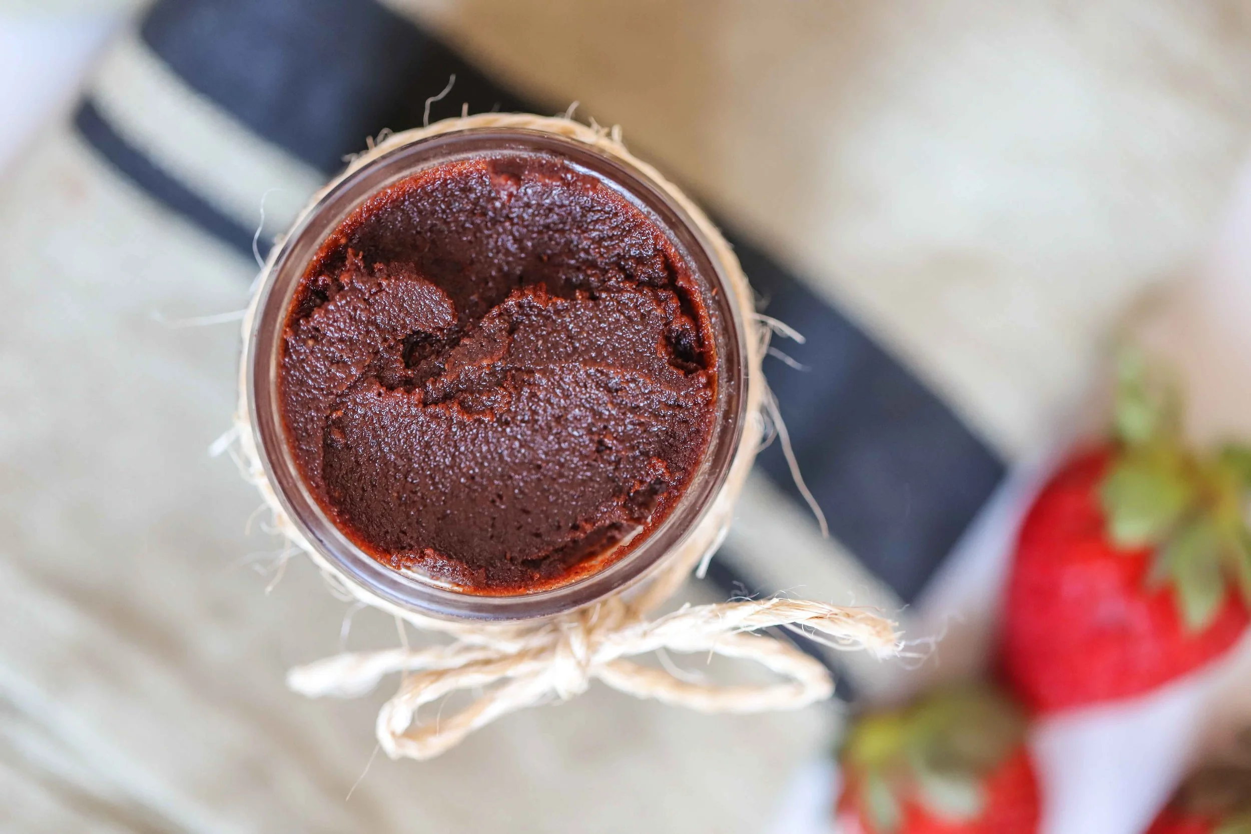 Close up of Glass jar of vegan nutella with twine wrapped around it in a bow on top of a linen napkin with a black stripe next to strawberries