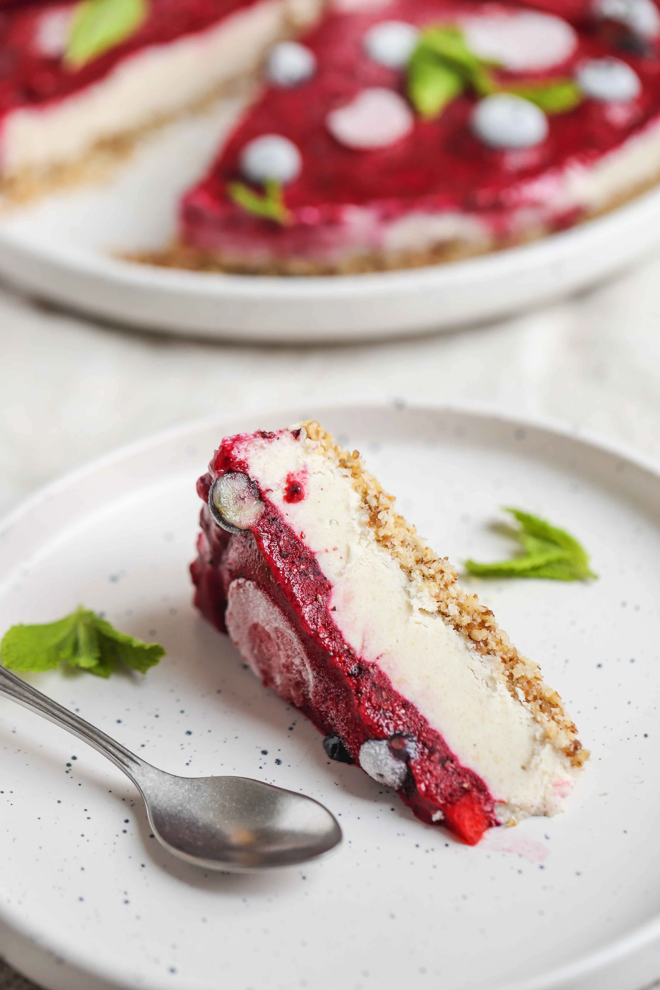 Slice of lemon berry cheesecake on a white plate with the whole cheesecake behind it