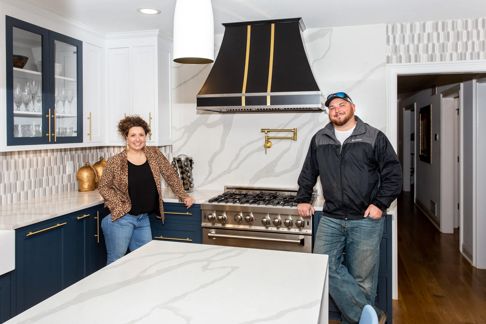 Betsy Manley with a renovation partner in a remodeled kitchen featuring navy cabinetry, quartz surfaces, brass fixtures, and a statement range hood.
