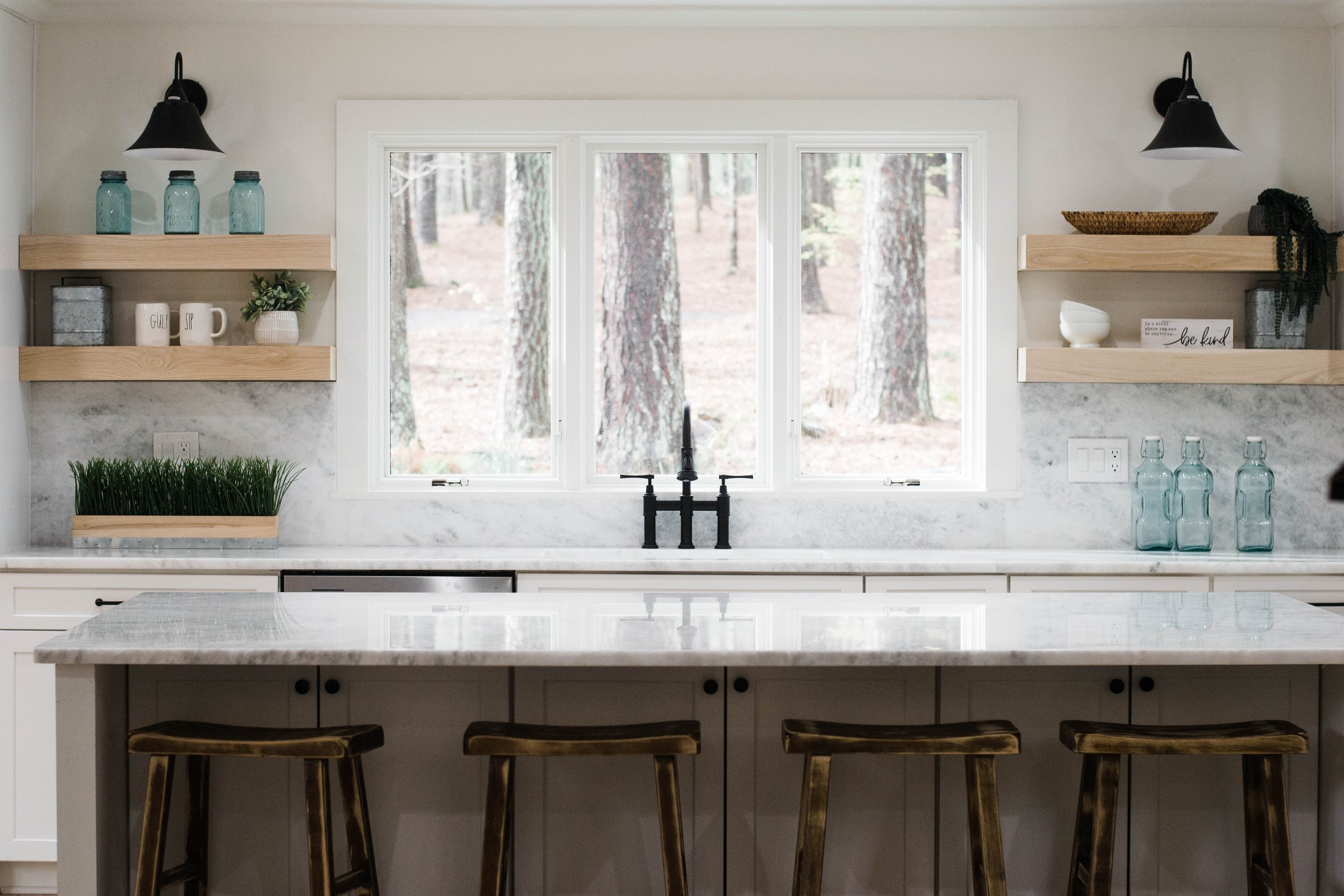 Bright renovated Lake Allatoona kitchen with white cabinetry, marble countertops, open wood shelving, black fixtures, and large windows overlooking the trees.