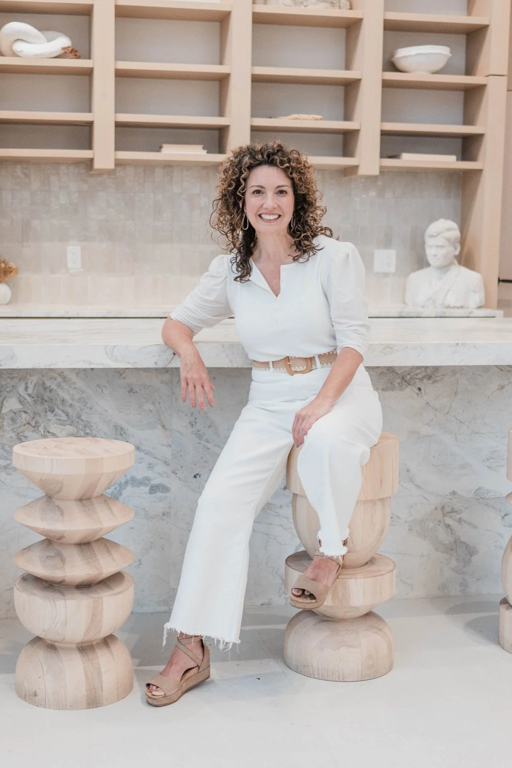 Betsy Manley, luxury interior designer and renovation expert, seated in a custom kitchen featuring marble surfaces and sculptural natural wood details.