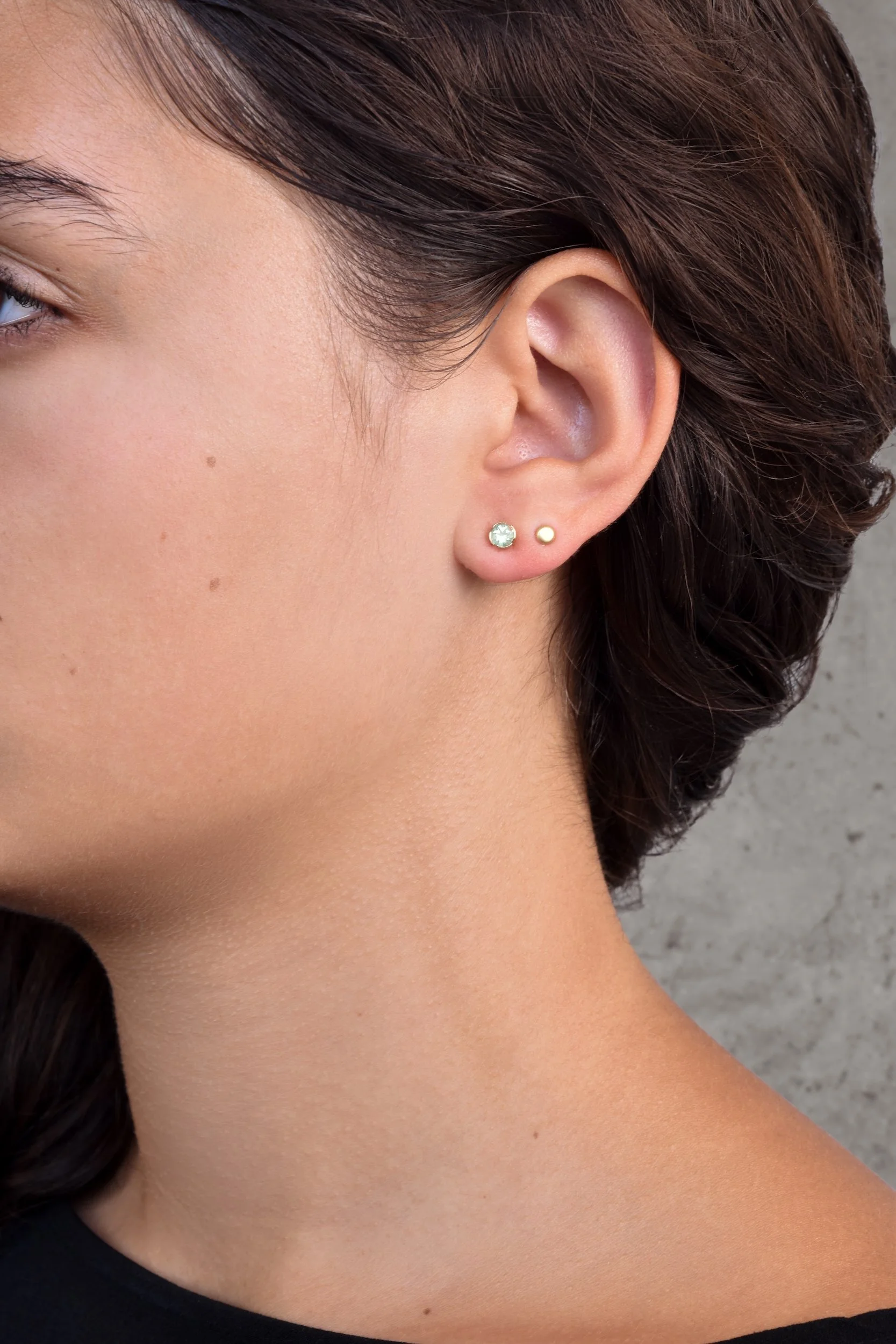 Elin Post Earrings — small gold studs with soft seafoam tourmaline stones worn on a model's ear against a grey concrete background