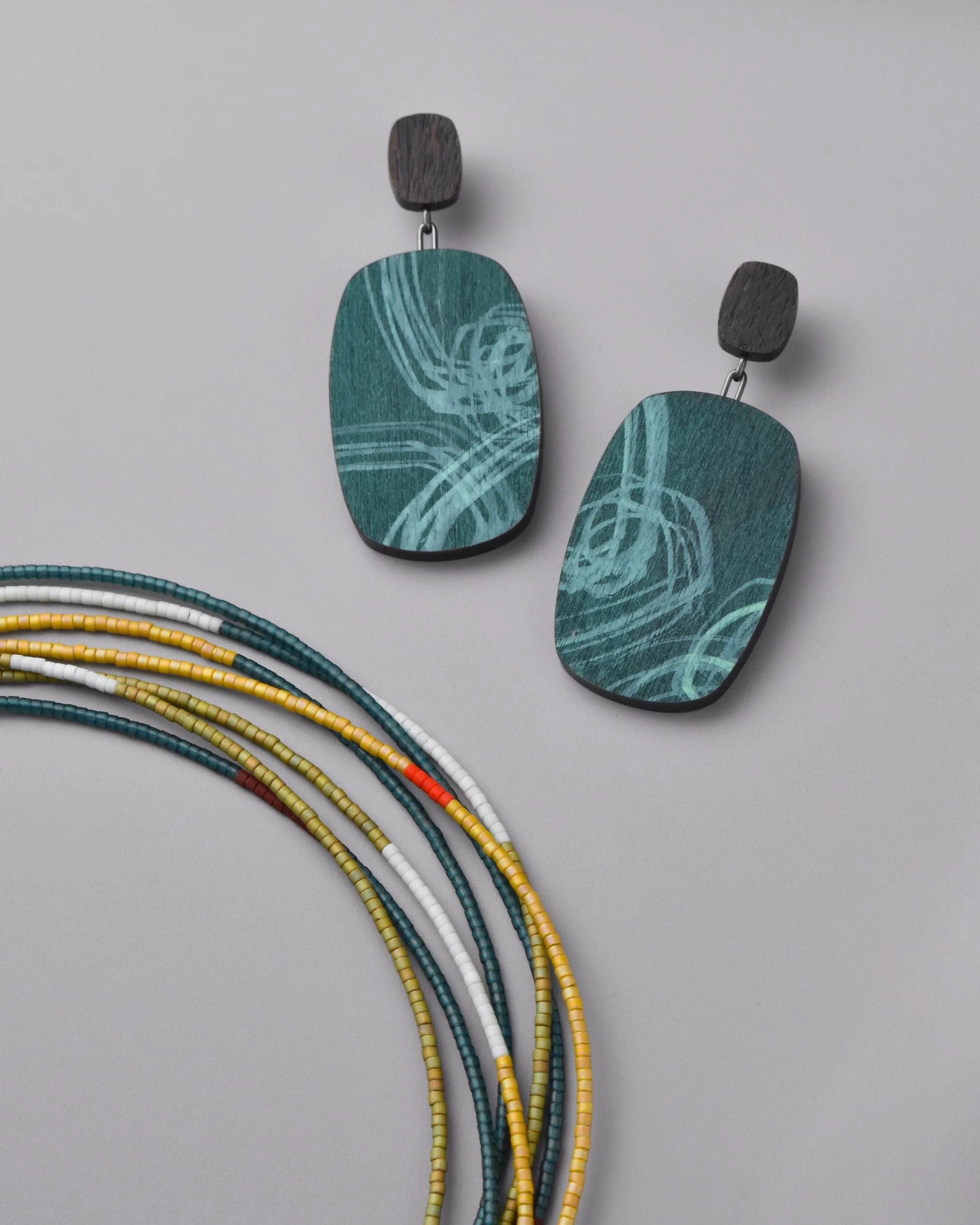 Audra Earrings — walnut and oxidized silver drop earrings at a jaunty angle on a grey background, showing the surging seafoam and teal ink layers over the natural wood grain