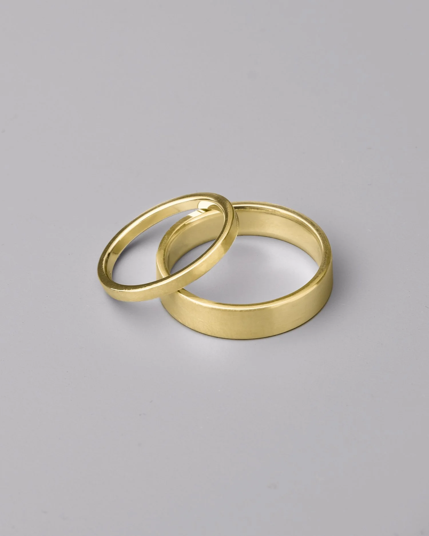 Two gold rings with a flat profile and satin finish lay stacked on a grey background.