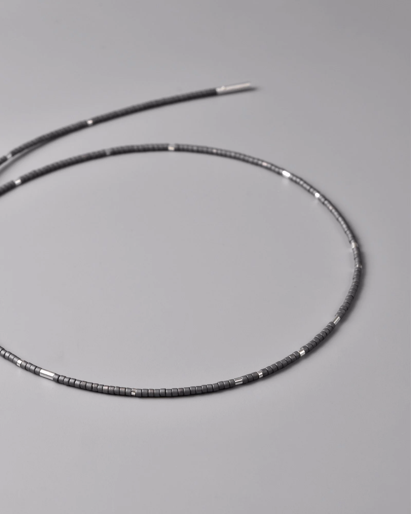 Clara Necklace — single-strand beaded necklace with charcoal grey Japanese glass beads and satin sterling silver tube segments, hanging vertically against a grey background