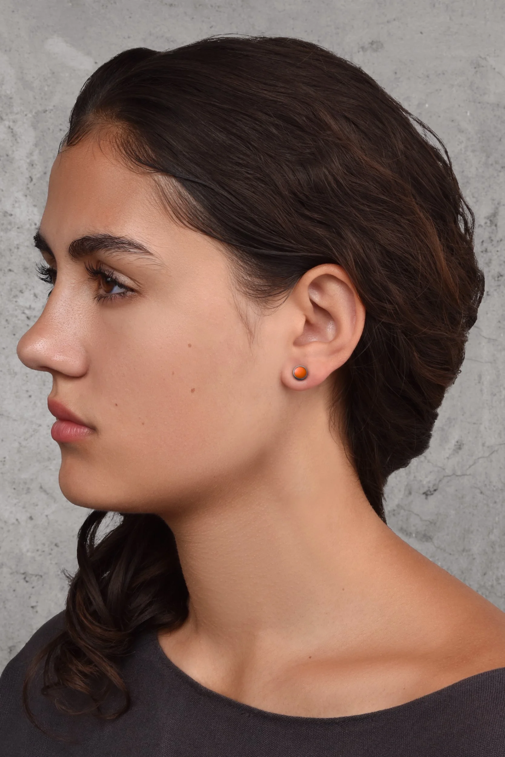 Frances Post Earrings in Mango — small warm mango orange enamel dot studs worn on a model's ear against a grey concrete background