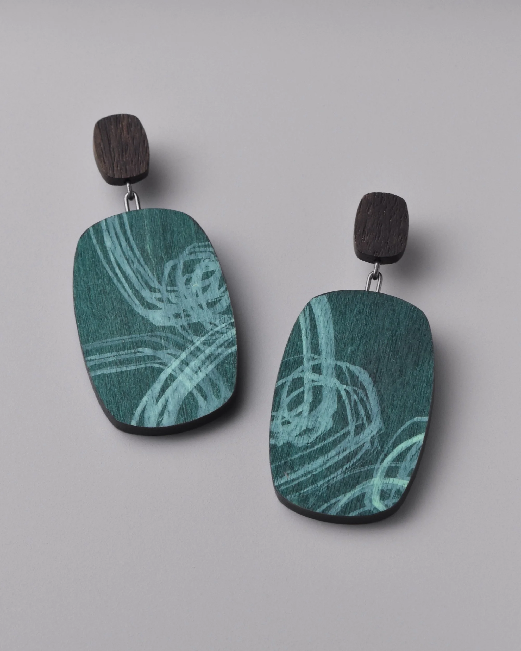 Audra Earrings — walnut and oxidized silver drop earrings at a jaunty angle on a grey background, showing the surging seafoam and teal ink layers over the natural wood grain