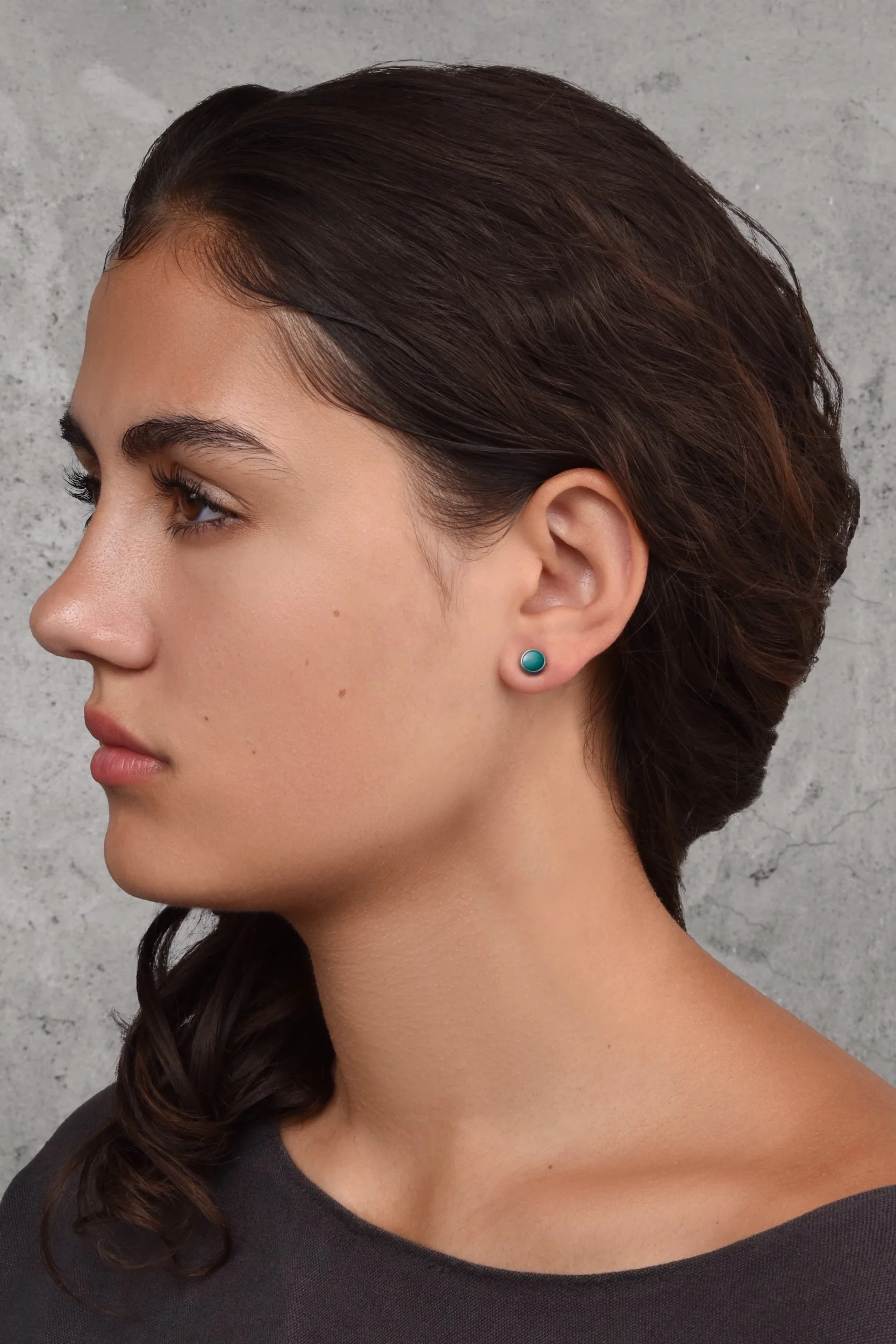 Frances Post Earrings in Lake — small soft lake blue enamel dot studs worn on a model's ear against a grey concrete background