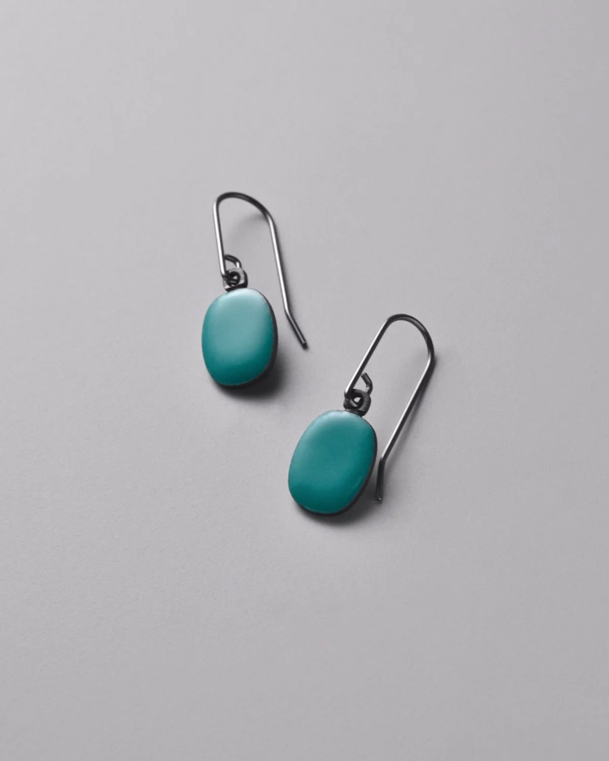 Emery Earrings in Lagoon — close-up detail of vivid turquoise enamel steel drop earrings on a grey background, showing the saturated color and small oval form