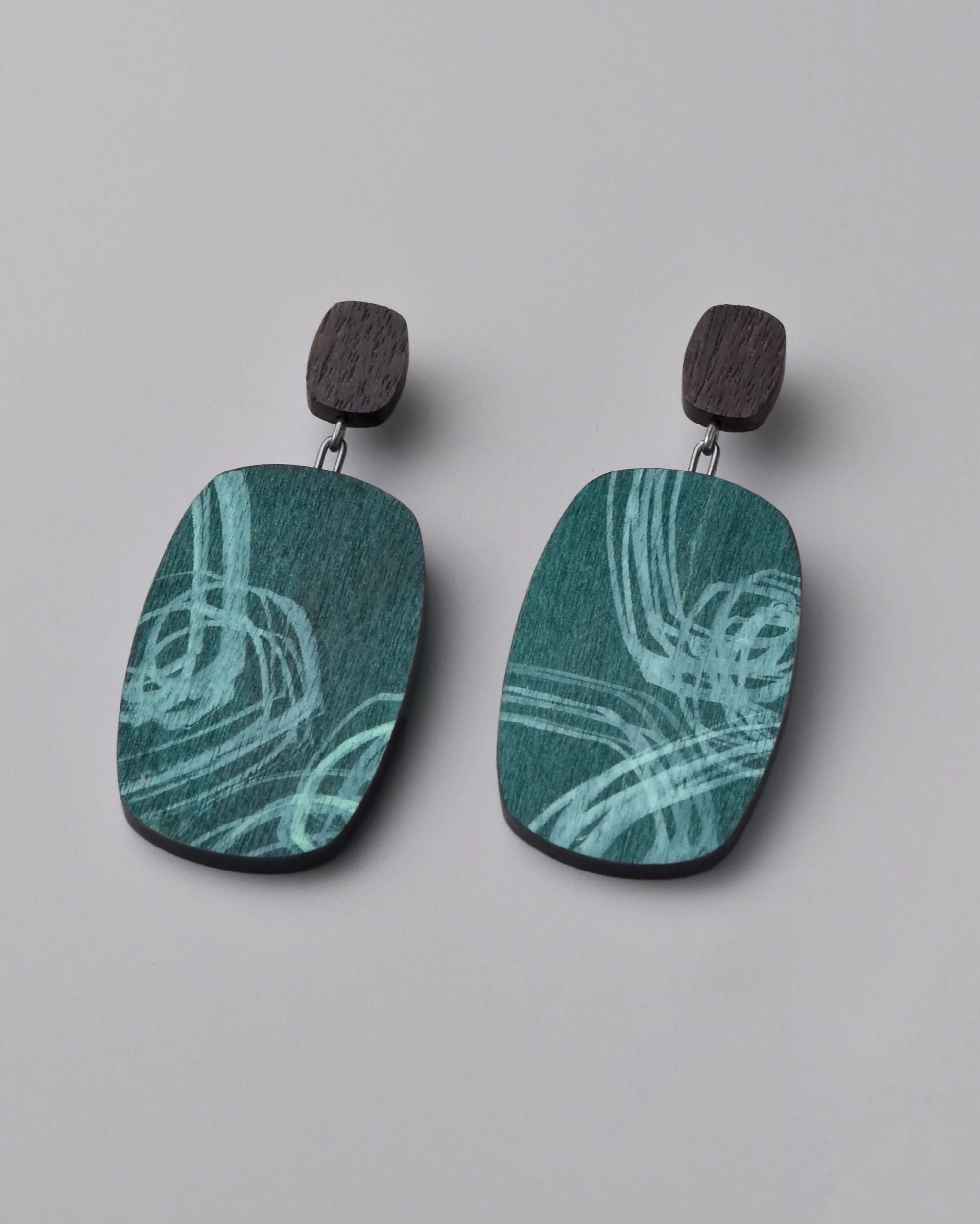 Audra Earrings — pair of drop earrings in oxidized sterling silver and walnut laying parallel on a grey background, with layers of seafoam and stormy teal ink