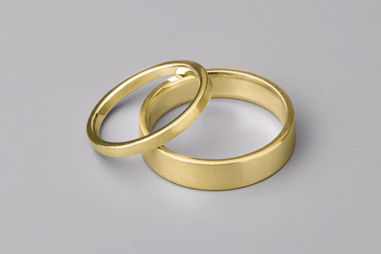 Two gold rings with a flat profile and satin finish lay stacked on a grey background