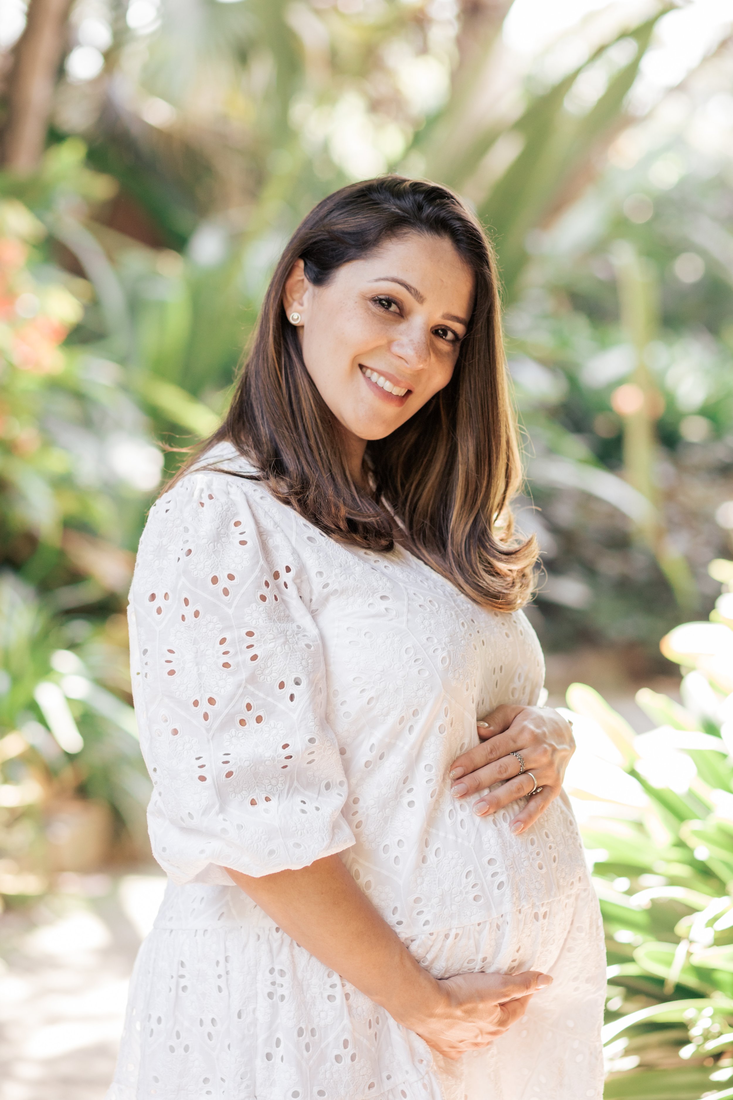 A pregnant woman with long brown hair, smiling, standing outdoors in a green garden, wearing a white embroidered dress. Stylish maternity portraits in Melbourne Gardens By Holly Rose Photography