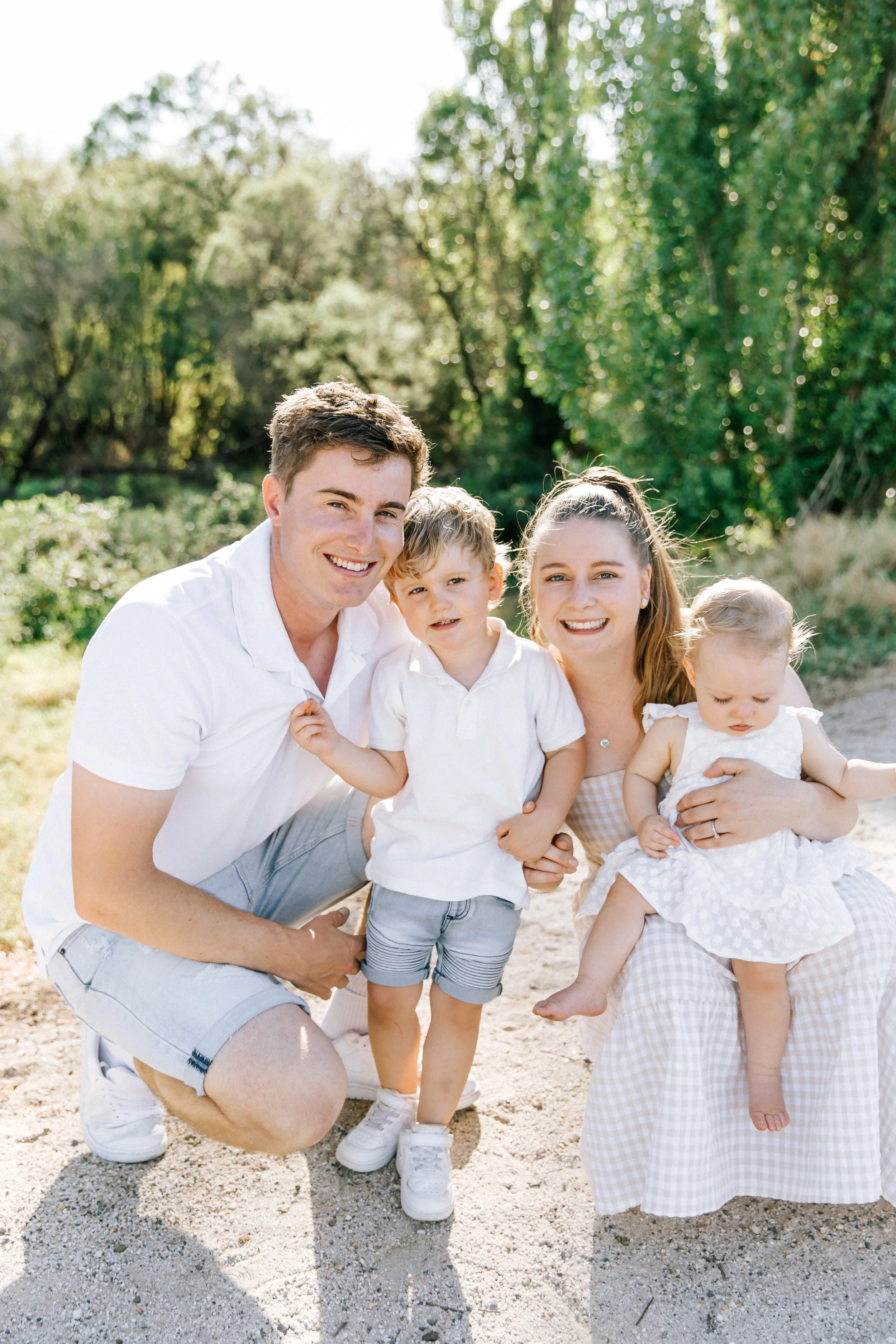 A family of five outdoors on a sunny day, smiling and posing together, with trees and greenery in the background. Elegant fine art family portraits By Holly Rose Photography
