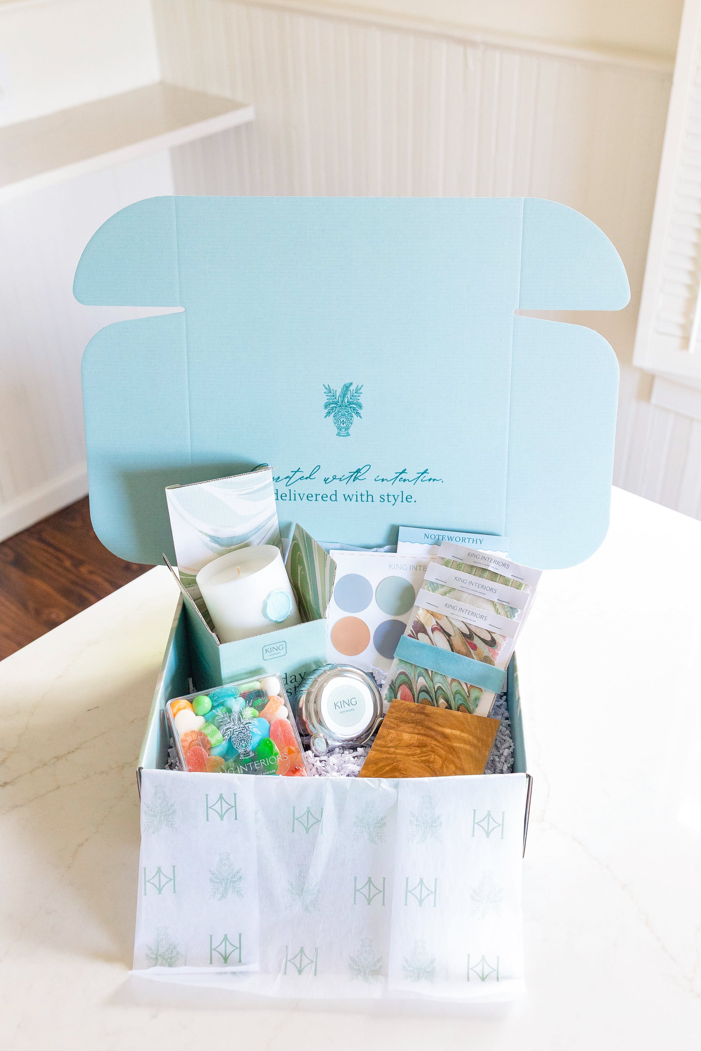Open teal box with King Interior branding on white table containing various items including a candle, candies, brochures, and promotional materials for King Interiors Curated Virutal Design services.