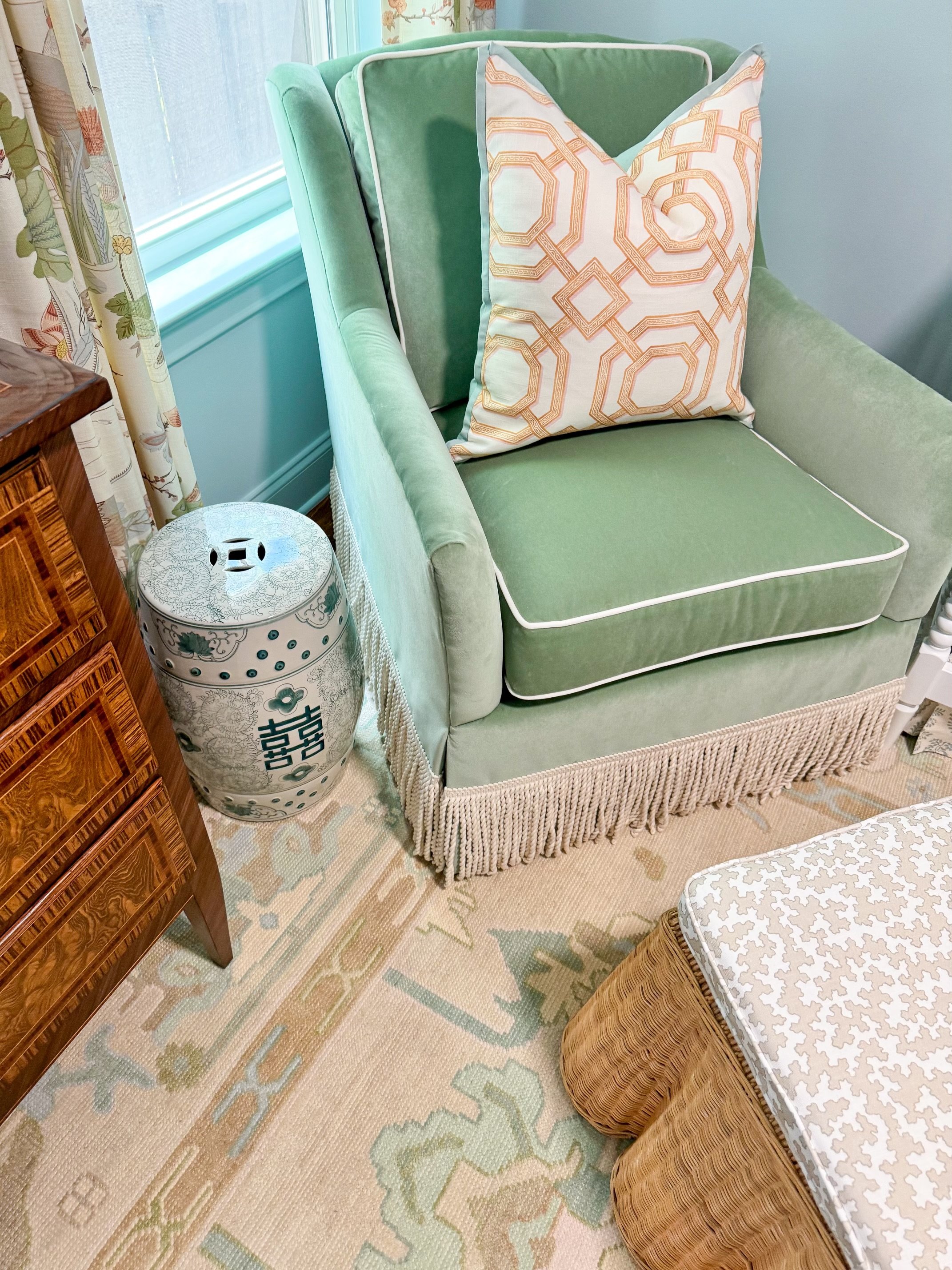 A green upholstered armchair with a decorative fringe at the base, featuring a patterned pillow. Next to it is a white ceramic stool with floral and Chinese characters, a wooden side table, a curtain with floral design, and patterned curtains by a wi