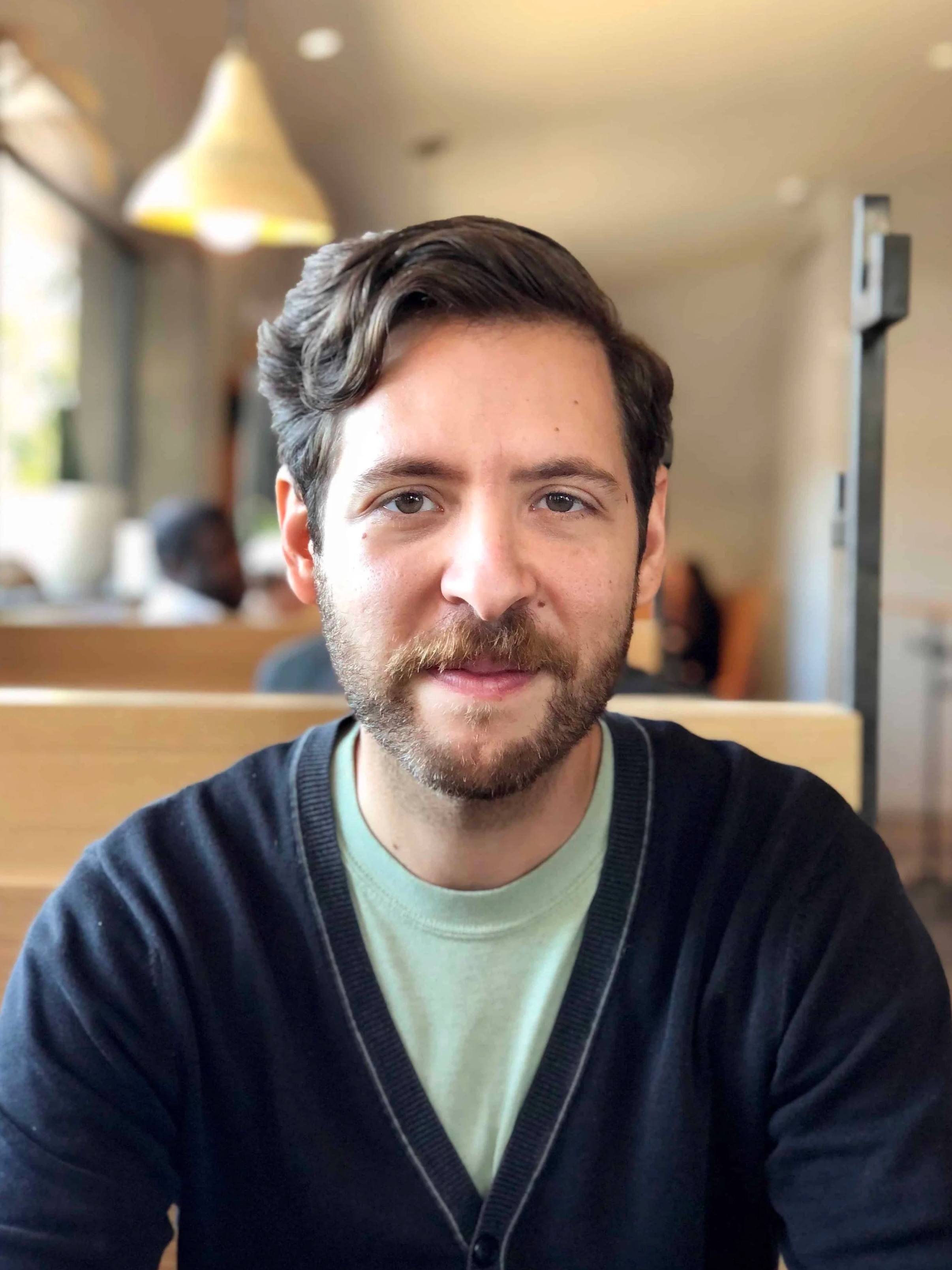 Alexander Lohn, designer and maker of custom wooden furniture for Alessandro & Maria Design. A young man with brown hair and a beard sitting at a table in a cafe, smiling at the camera, with a blurred background of other people and cafe decor.
