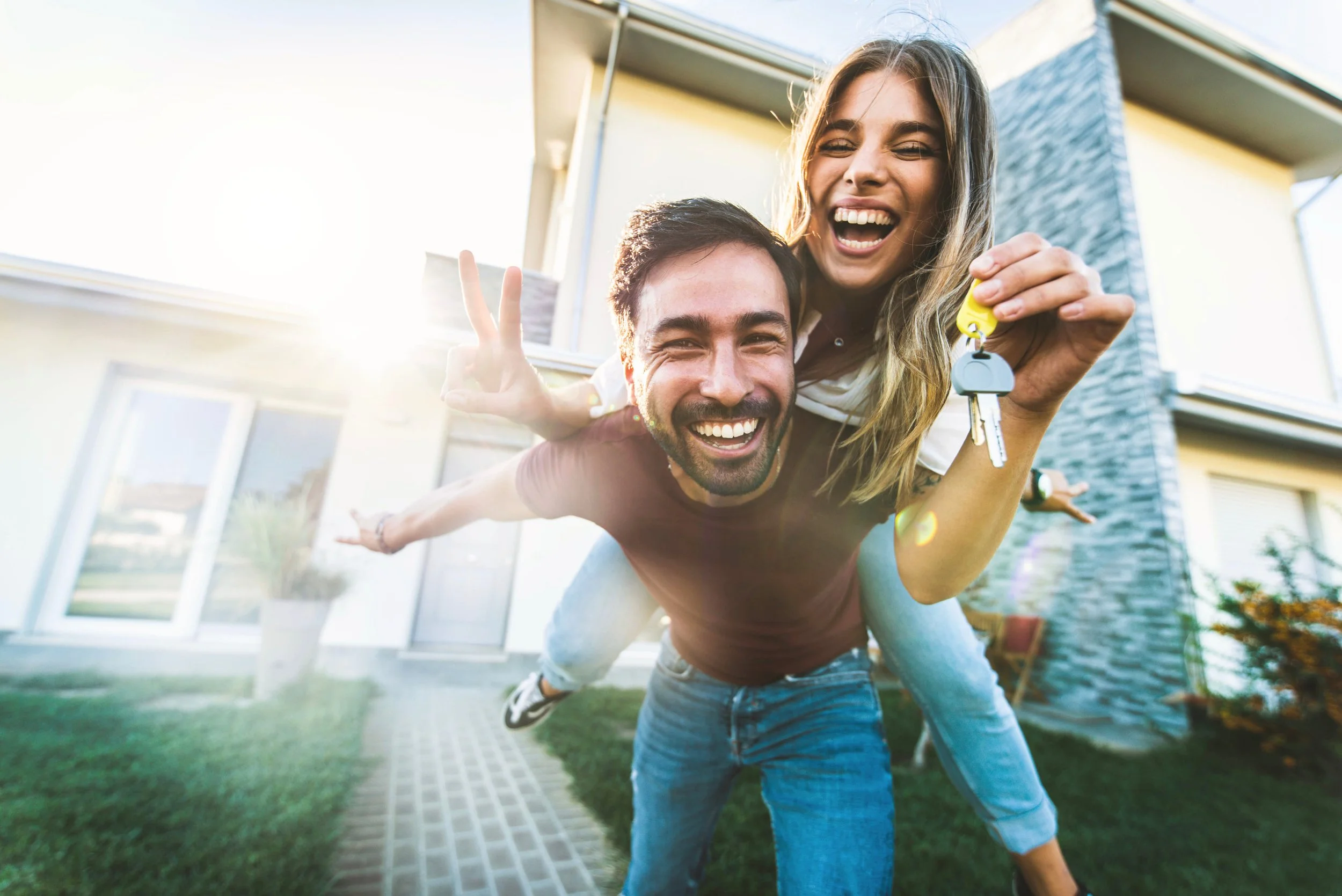 A smiling young woman sitting on the man's back, holding a key, with a happy couple standing outside their modern house during sunlight.