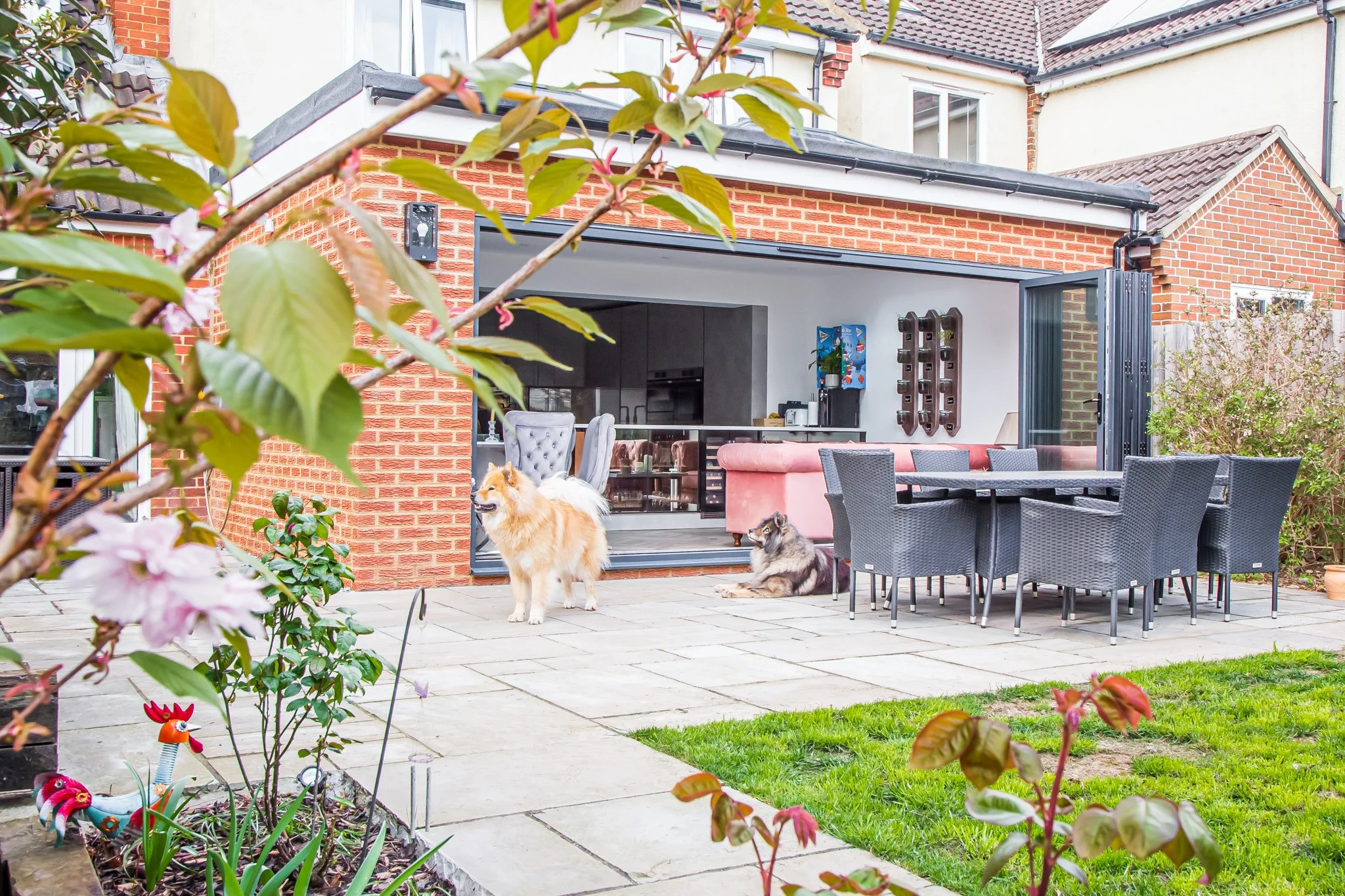 View of a backyard patio with a brick house in the background. There is a dining table with eight black wicker chairs on a tiled patio. Two dogs, one small and fluffy and one larger with a thick coat, are present, with the larger dog standing and the smaller dog lying down. There are plants and shrubs in the foreground, and an open sliding glass door leads into the house, revealing a modern interior with a pink sofa.