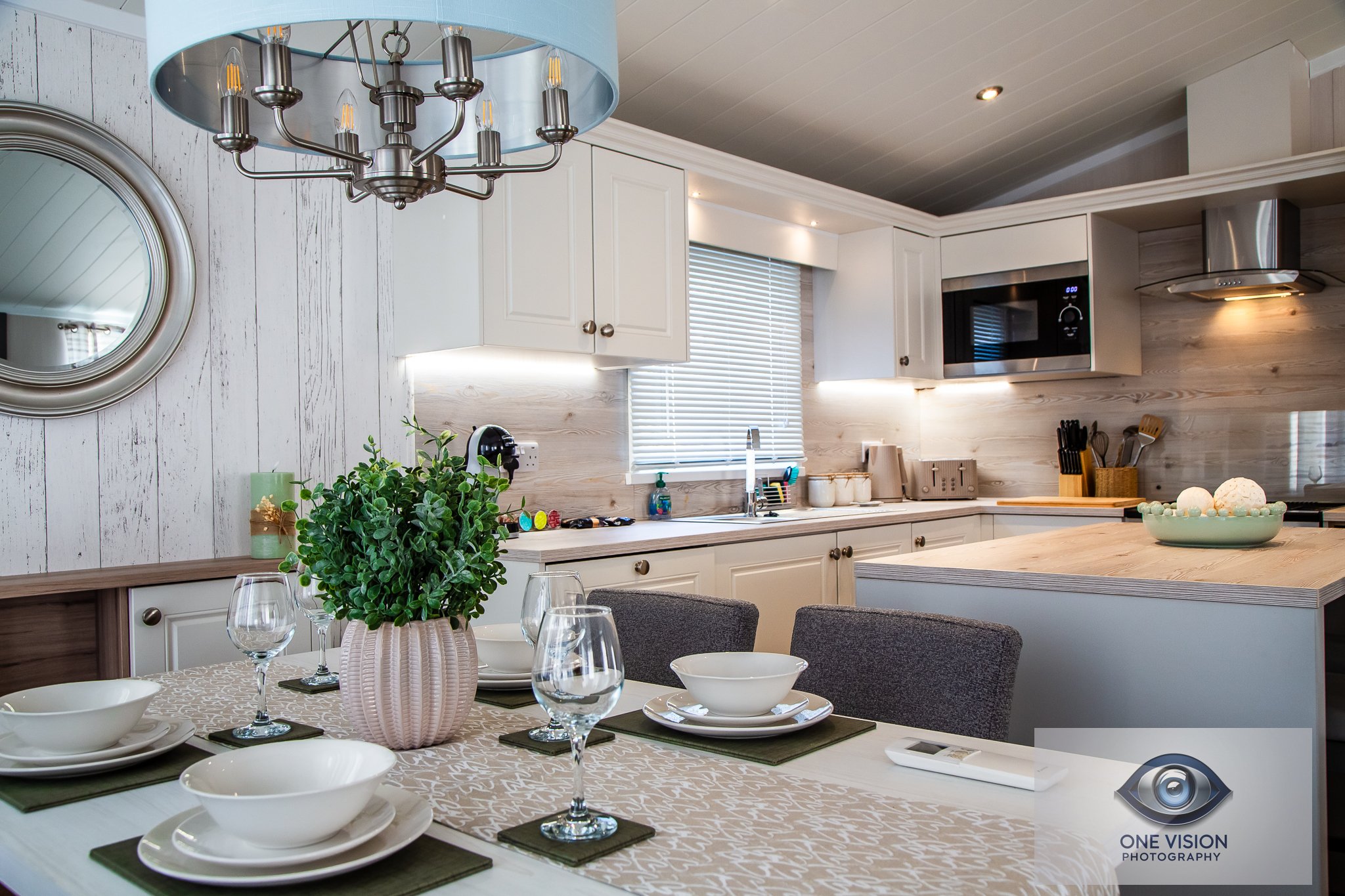 A modern kitchen and dining area with a table set for four, decorated with a potted green plant, white dinnerware, glasses, and a beige table runner; the kitchen has white cabinets, a window with blinds, a microwave, and various kitchen utensils.