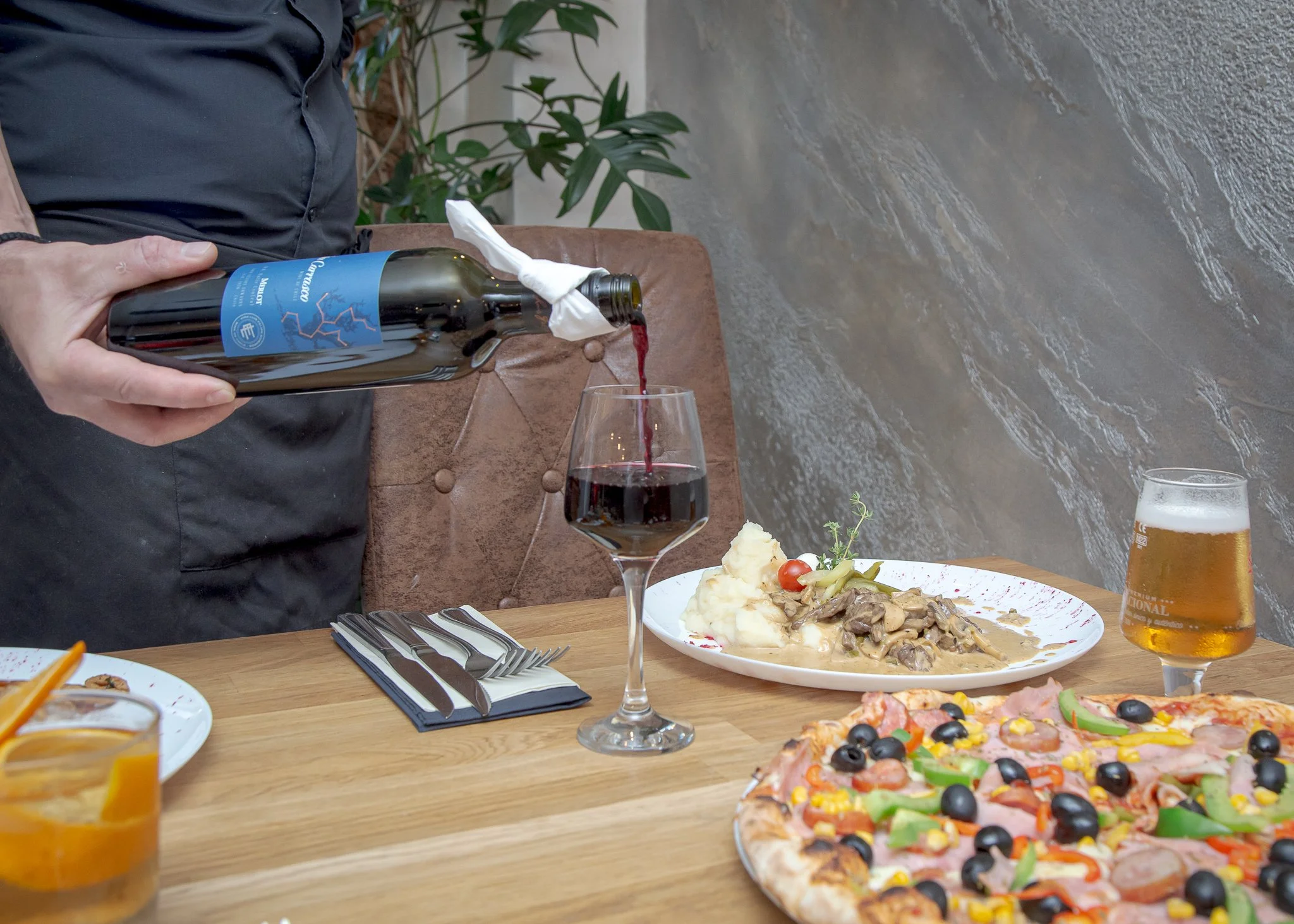 Person pouring red wine into a glass at a dining table with pizza, a plate of mashed potatoes and meat, and a glass of beer.
