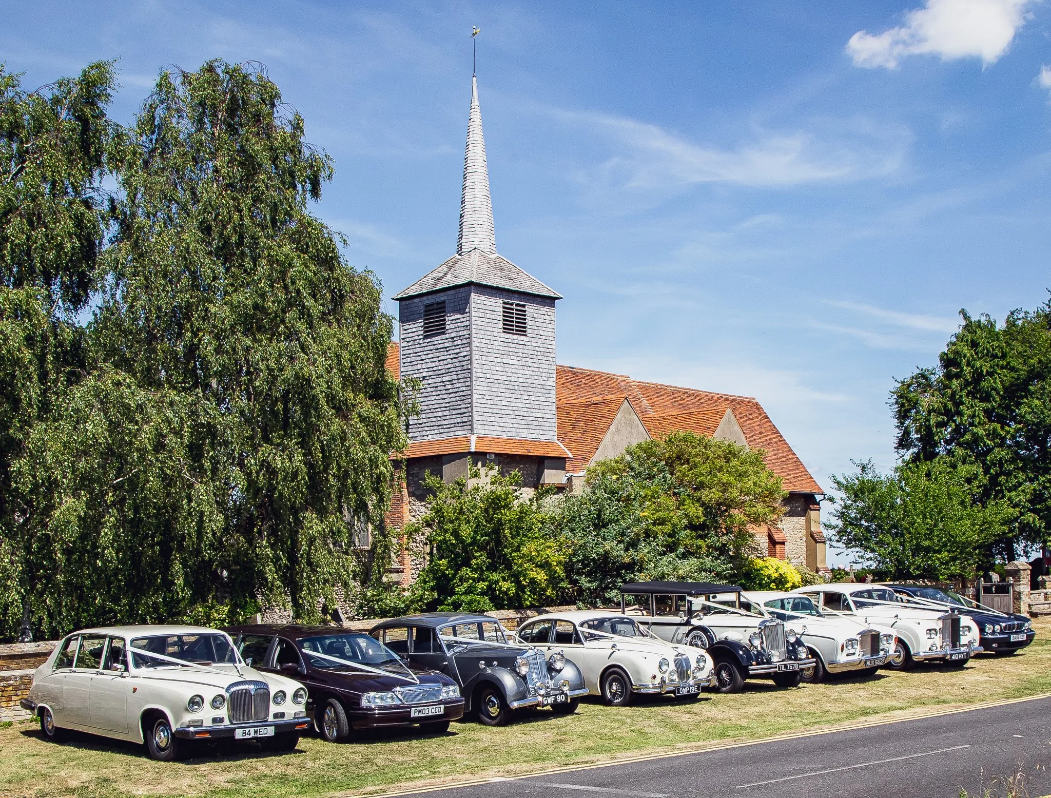 A row of vintage cars parked on grass outside a church with a tall steeple, trees, and a blue sky.