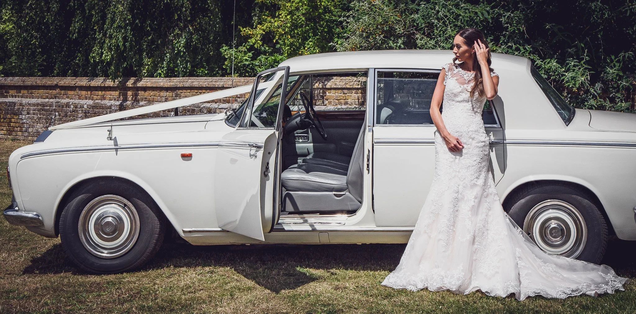 A bride in a white lace wedding dress standing outside next to a vintage white car with open doors, on a grassy area with trees and a brick wall in the background.