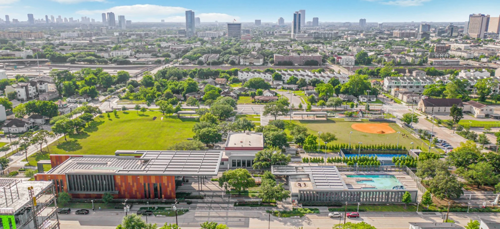 Aerial view of a city park with green grass, trees, a baseball field, and playgrounds, surrounded by residential areas and high-rise buildings in the background.