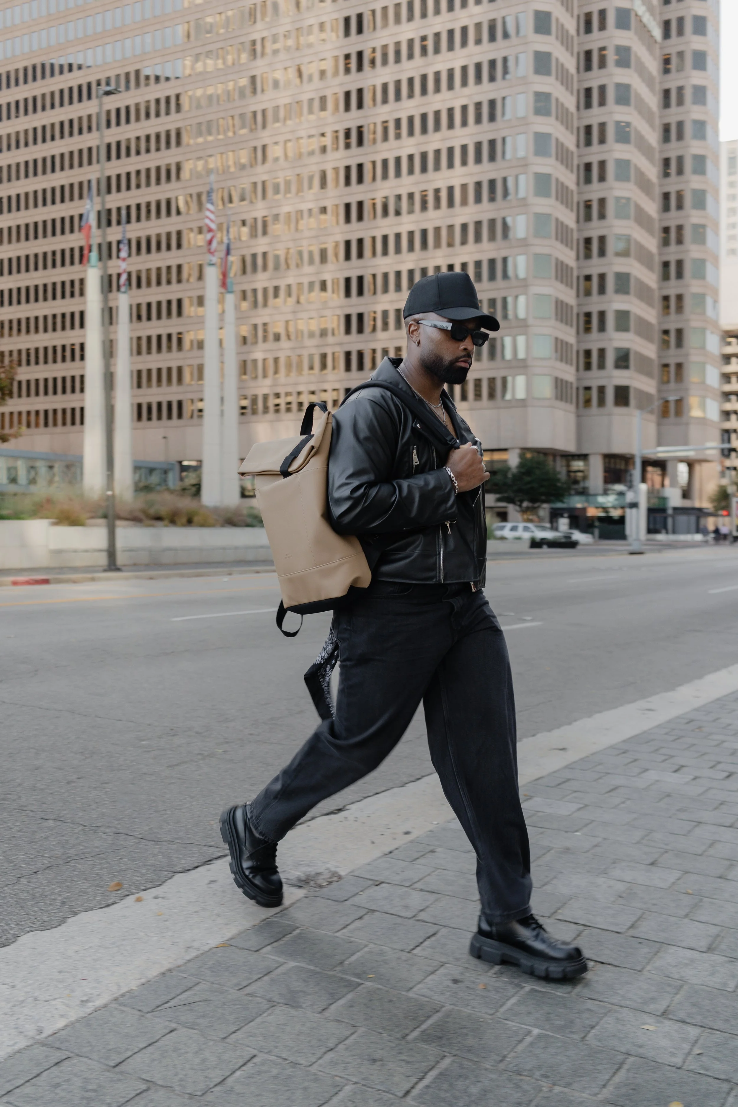 Man walking on city sidewalk wearing black leather jacket, black pants, black boots, black cap, sunglasses, and carrying a beige backpack in front of tall office buildings.